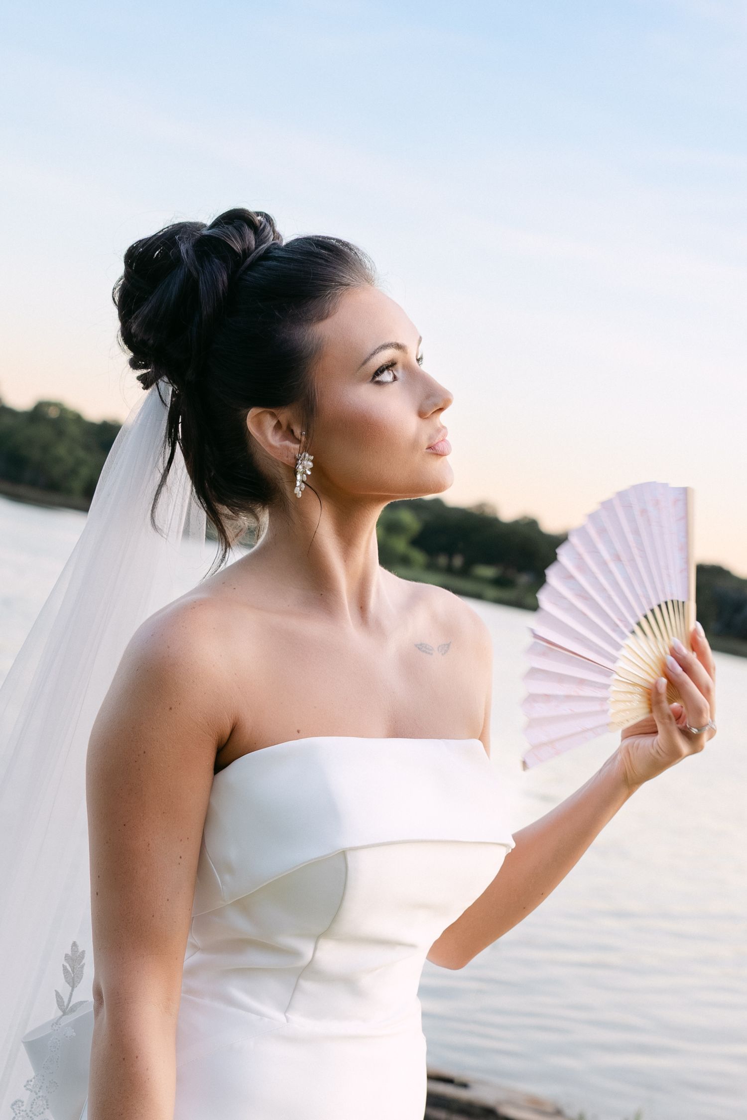 A bride in a strapless white dress poses gracefully with a delicate paper fan by the water at sunset.