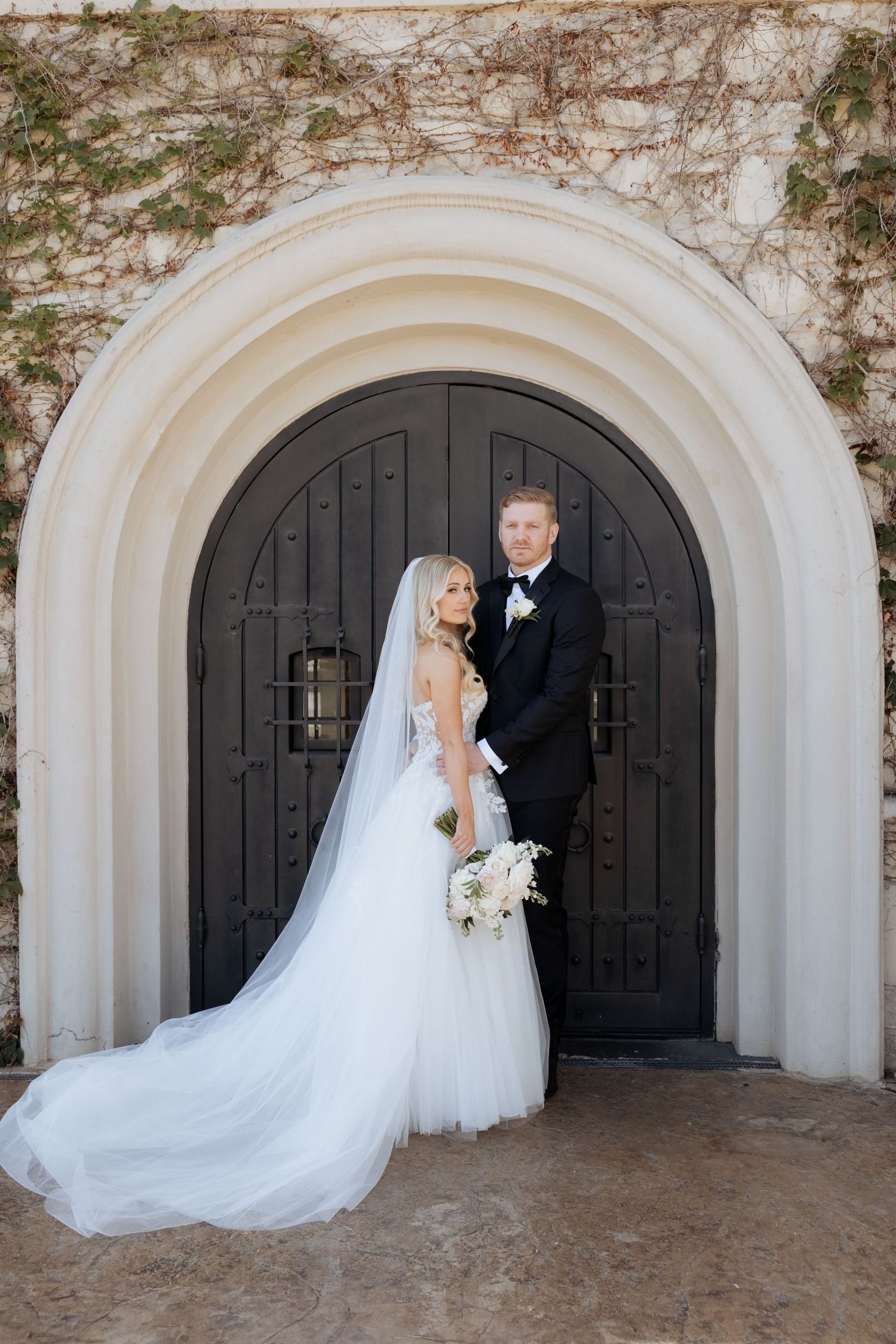 Bride and groom posing elegantly before a grand arched black door, bride in a flowing white gown with veil and bouquet.