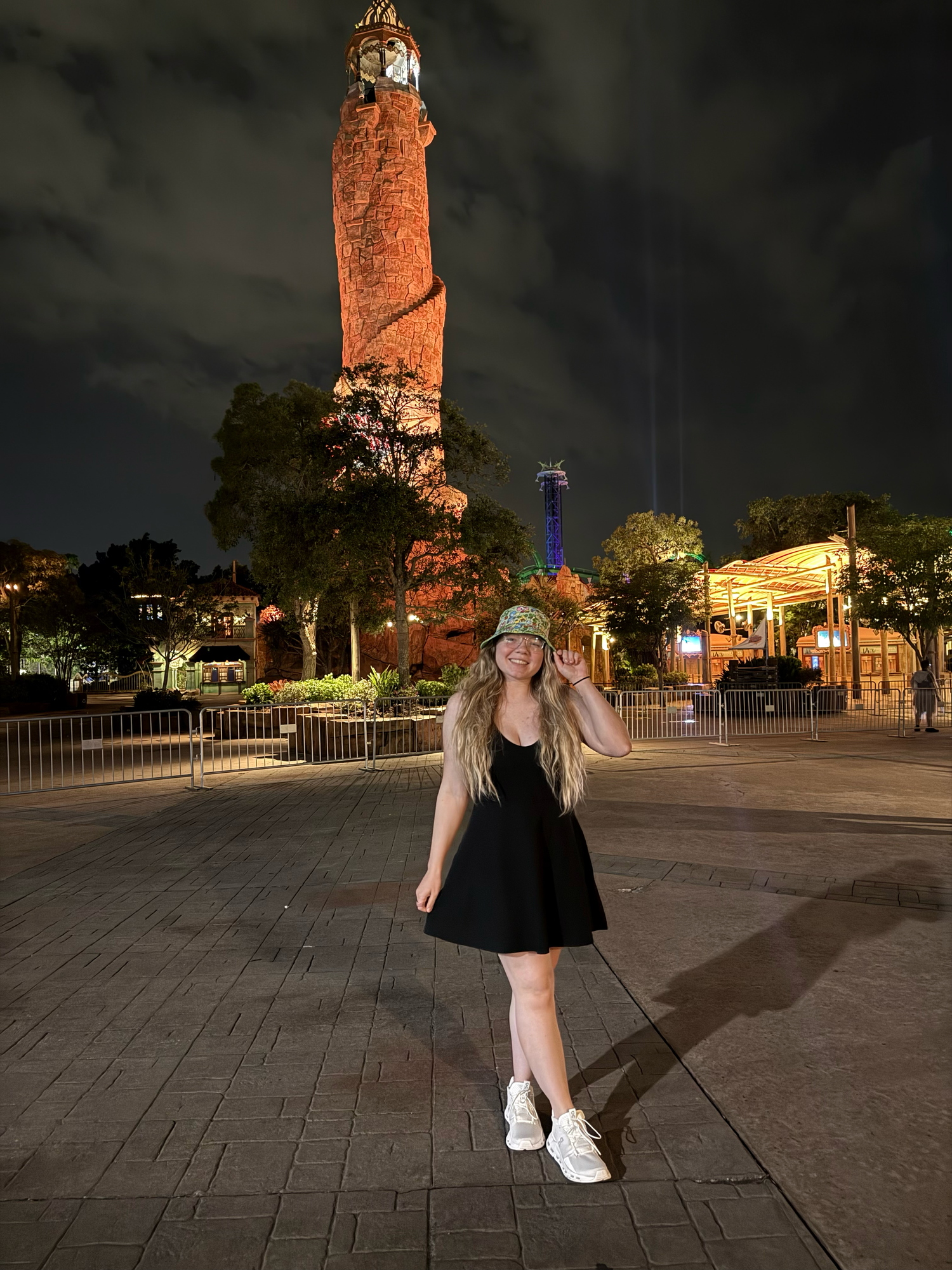 A person wearing a black dress and white sneakers poses against a dark nighttime cityscape with an illuminated tower.
