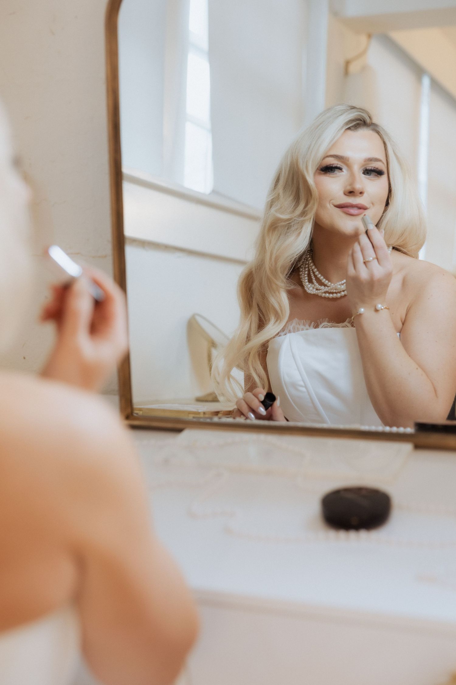 A bride in white preparing her makeup while looking in a mirror, wearing a necklace with a soft romantic aesthetic.
