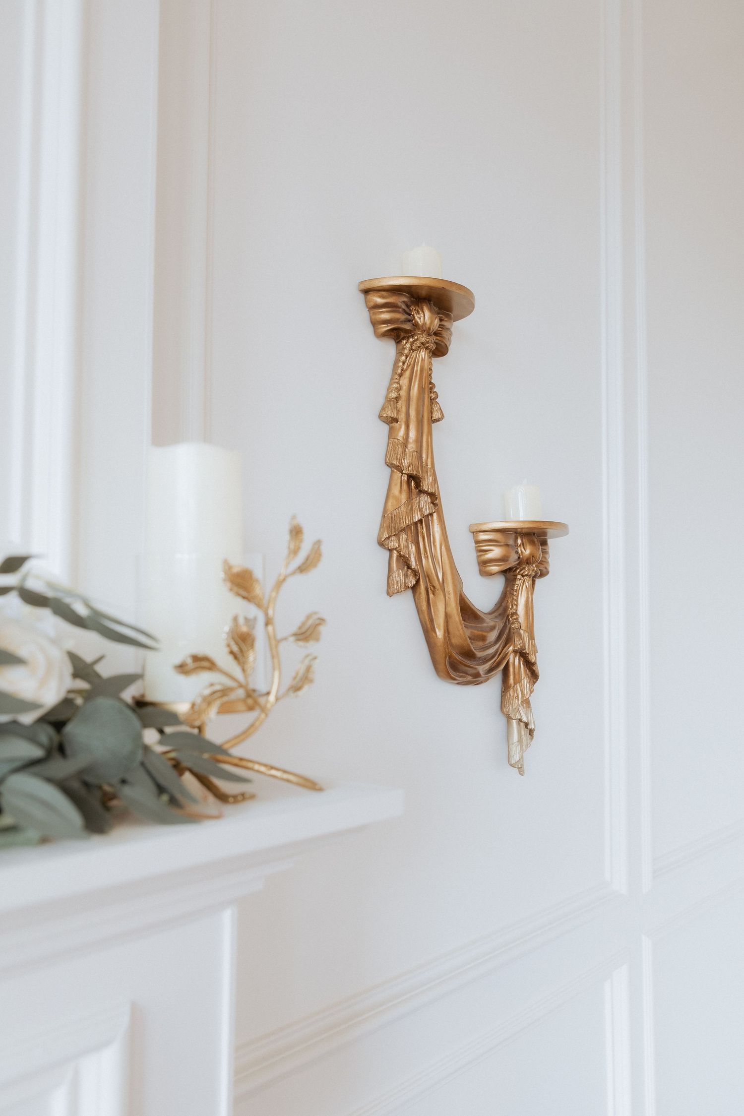 Gold decorative wall sconces with floral accents mounted on white paneled walls create an elegant vintage aesthetic.