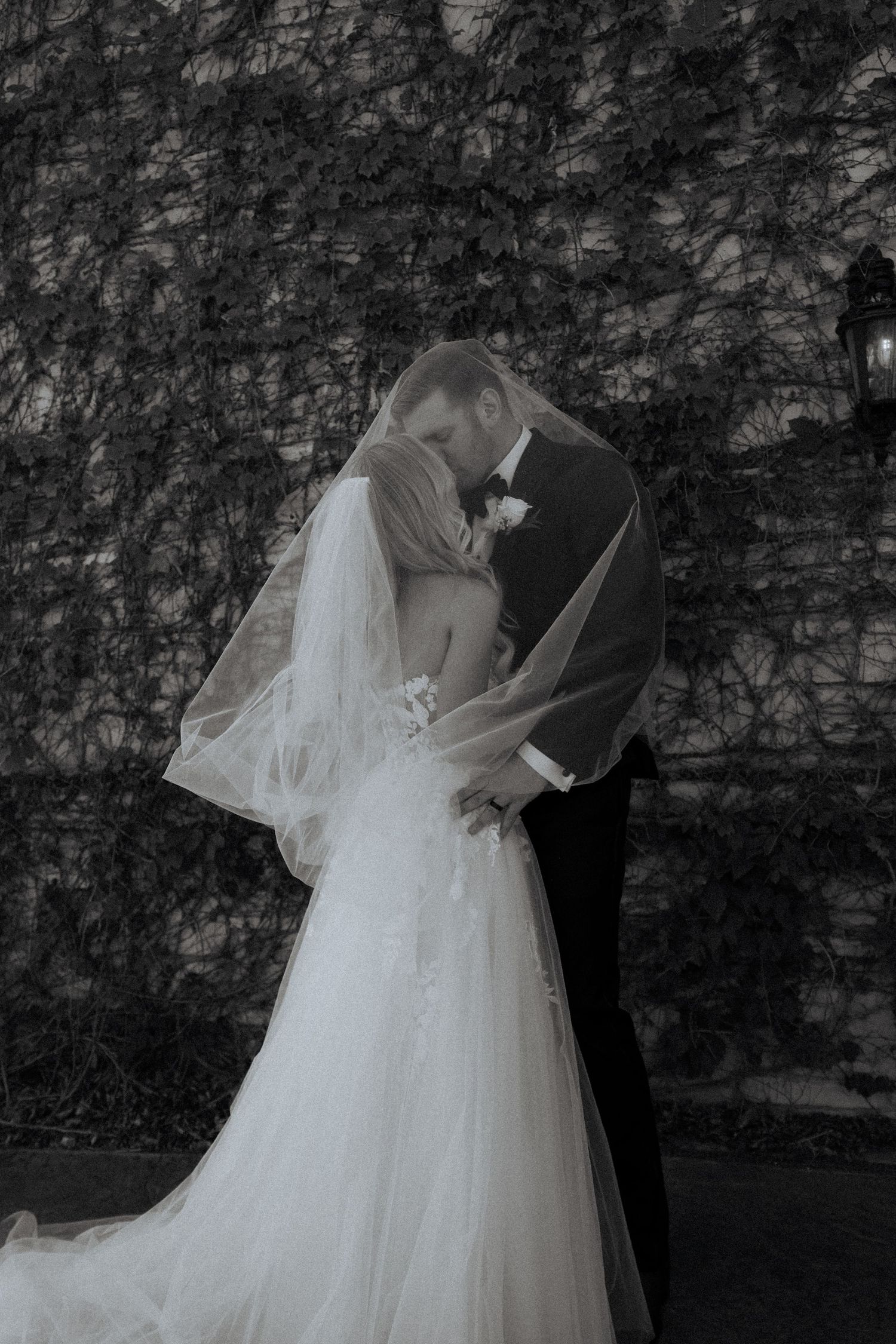 Black and white photo of bride and groom kissing under veil against ivy-covered wall.
