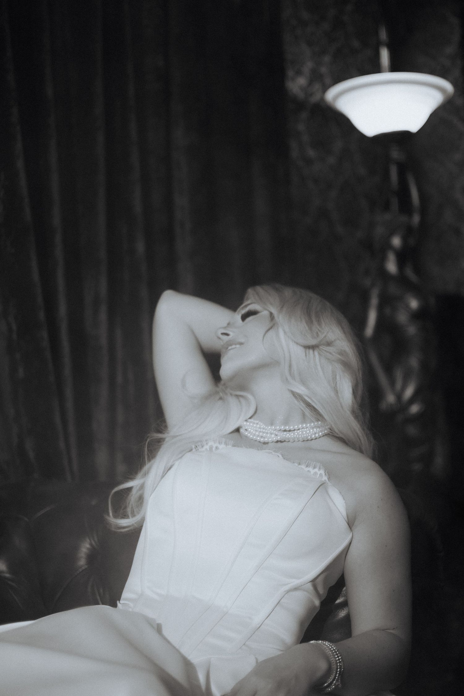 Black and white photo of a woman in a white dress reclining on a chair, with a pearl necklace and dramatic lighting.