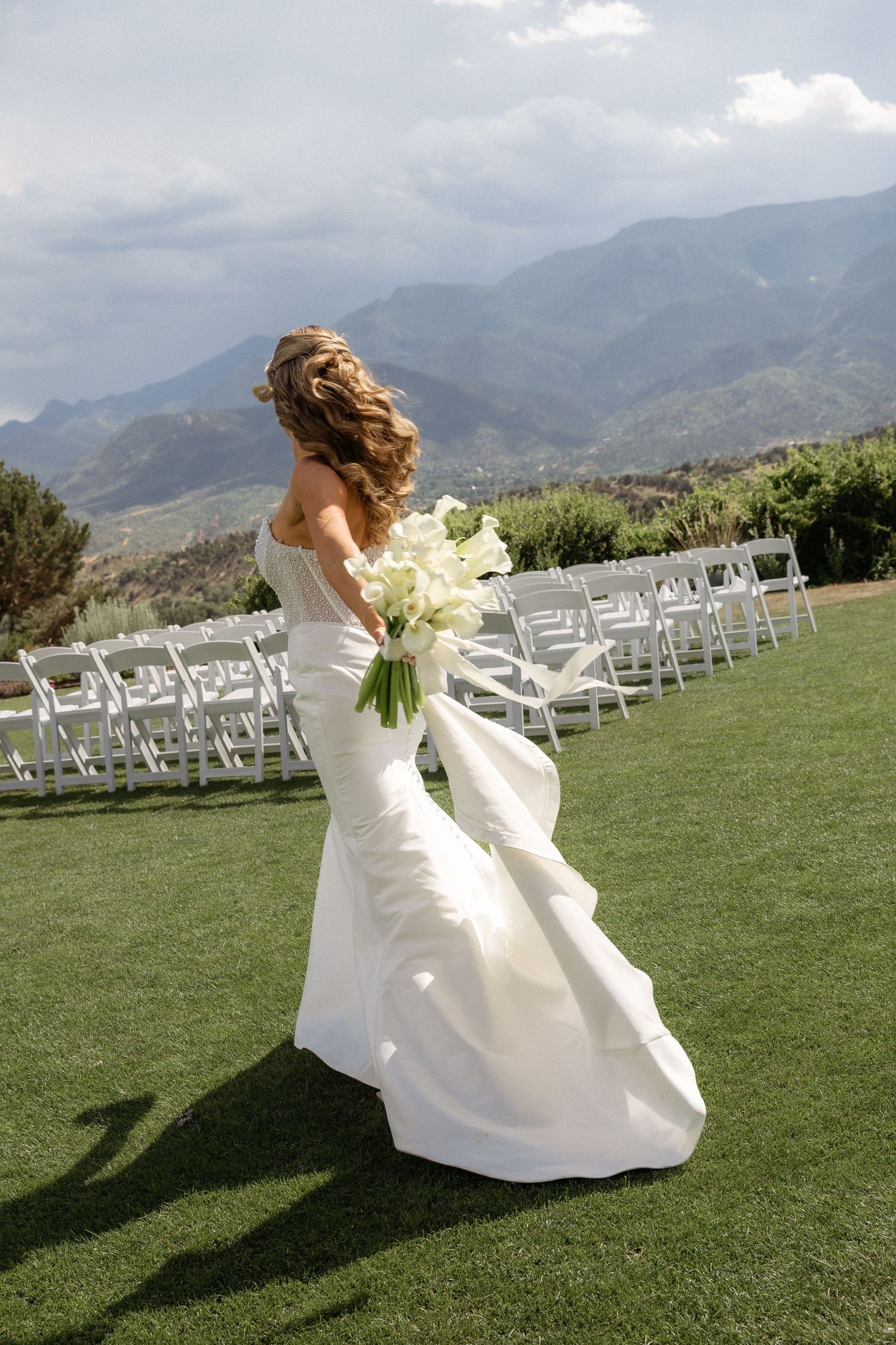 Bride in white mermaid gown holding bouquet of white flowers, standing on green lawn with mountain backdrop.