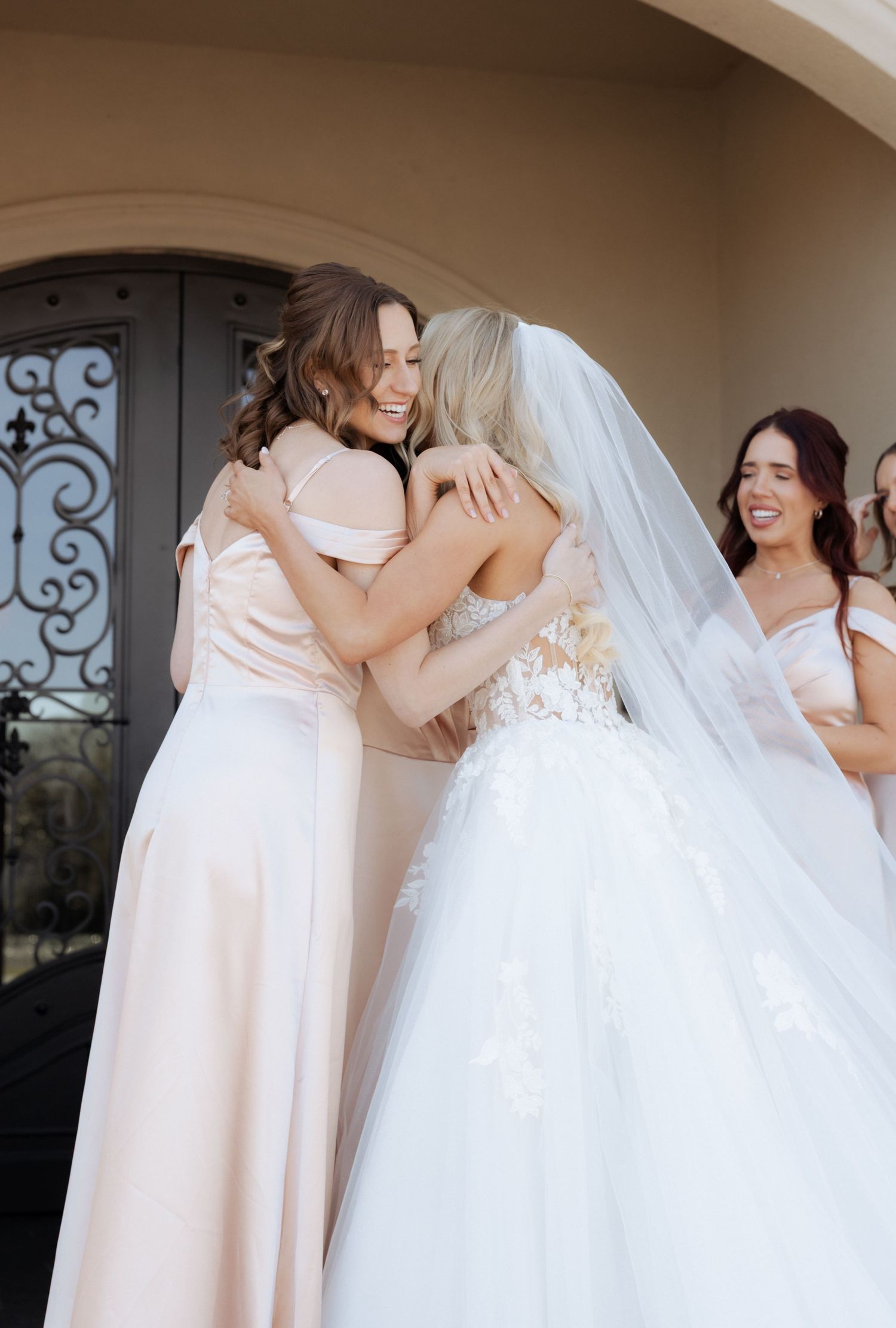 Bride in white lace gown and veil hugging bridesmaid in blush pink dress outside elegant venue.