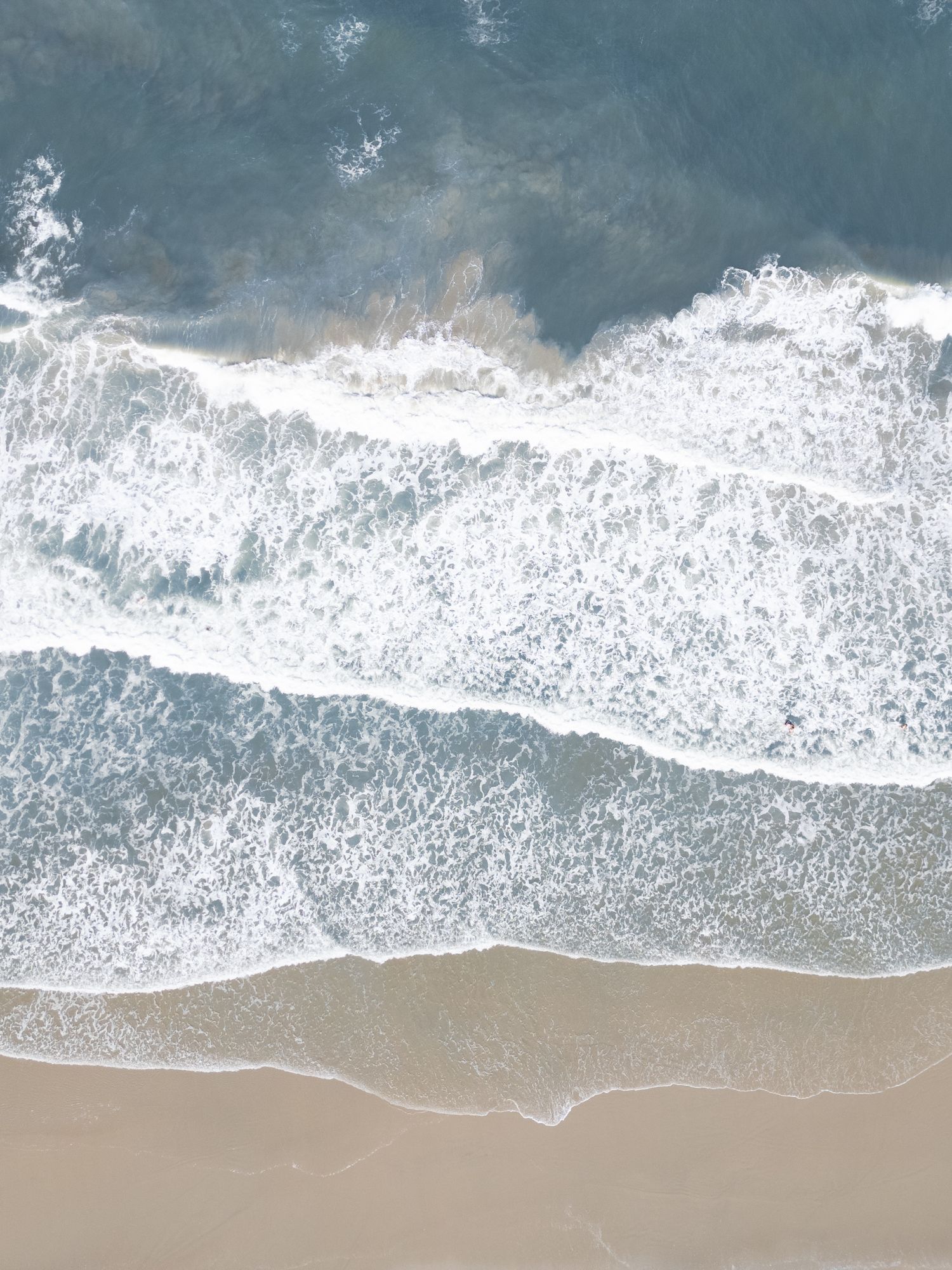 Aerial view of white ocean waves crashing onto a sandy beige beach with turquoise water stretching to the horizon.