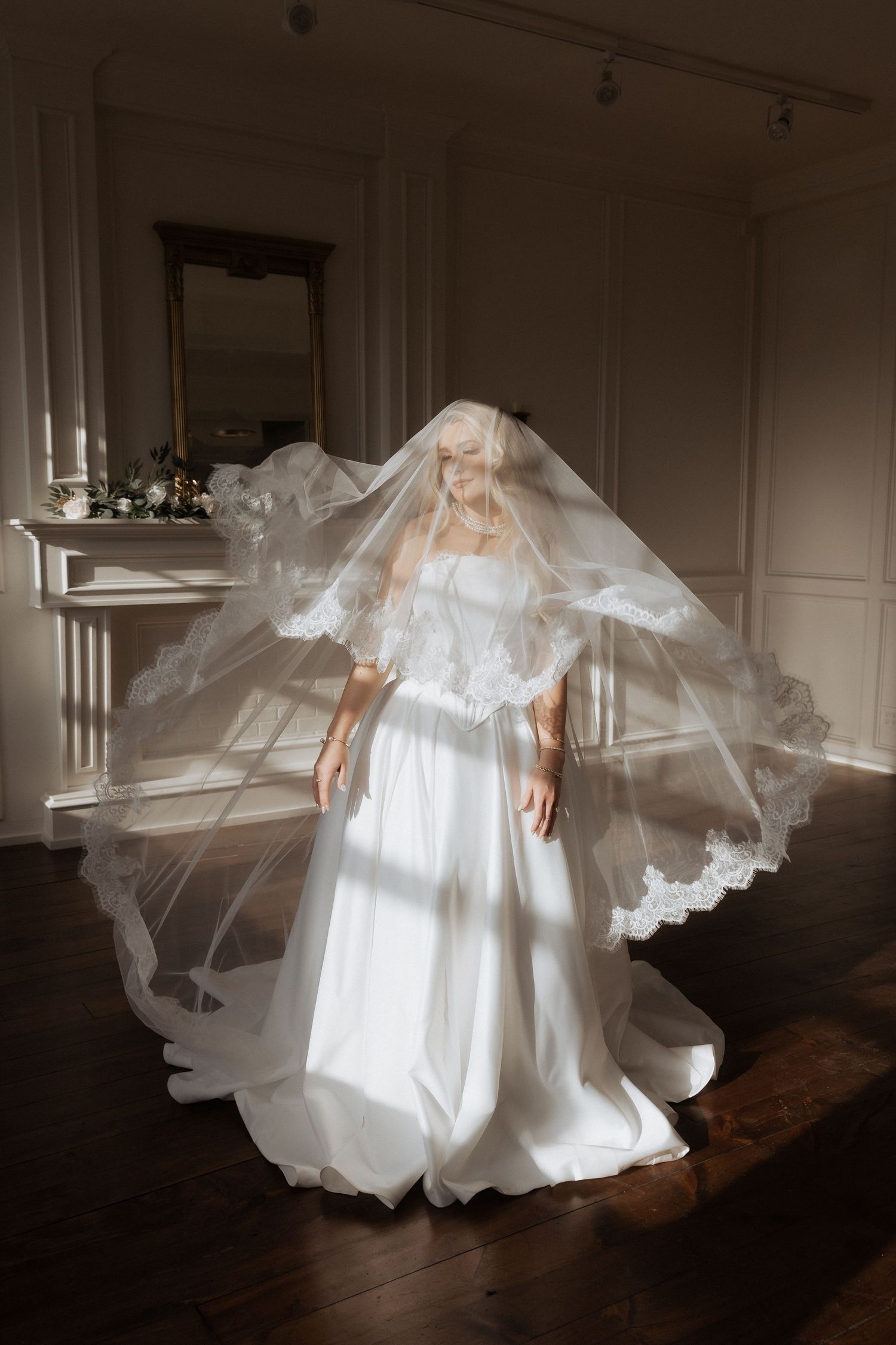 Flowing white wedding dress with dramatic billowing veil captured in moody indoor lighting against white wall and fireplace.