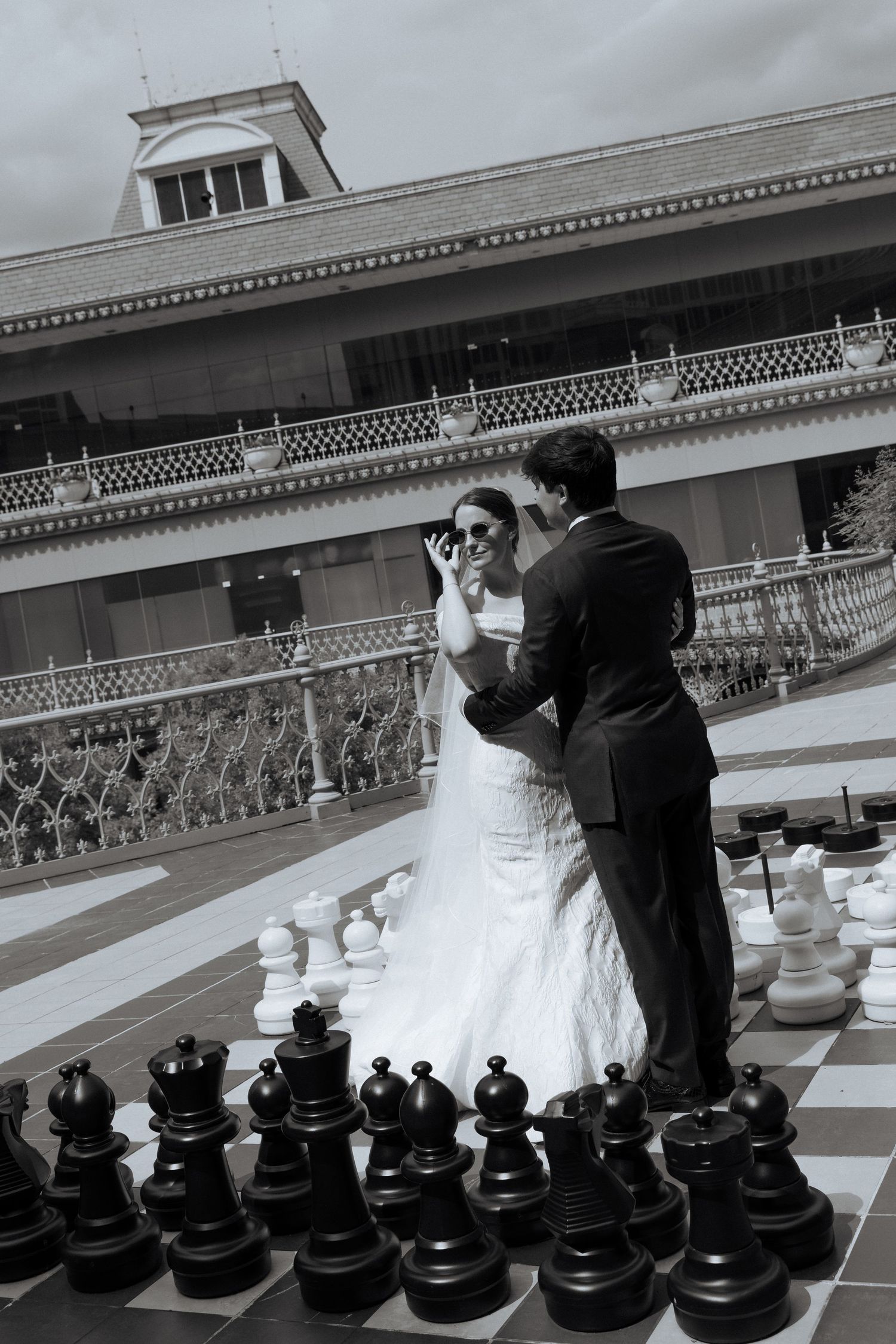 Black and white photo of a bride and groom standing on a giant chess board outside an elegant building.
