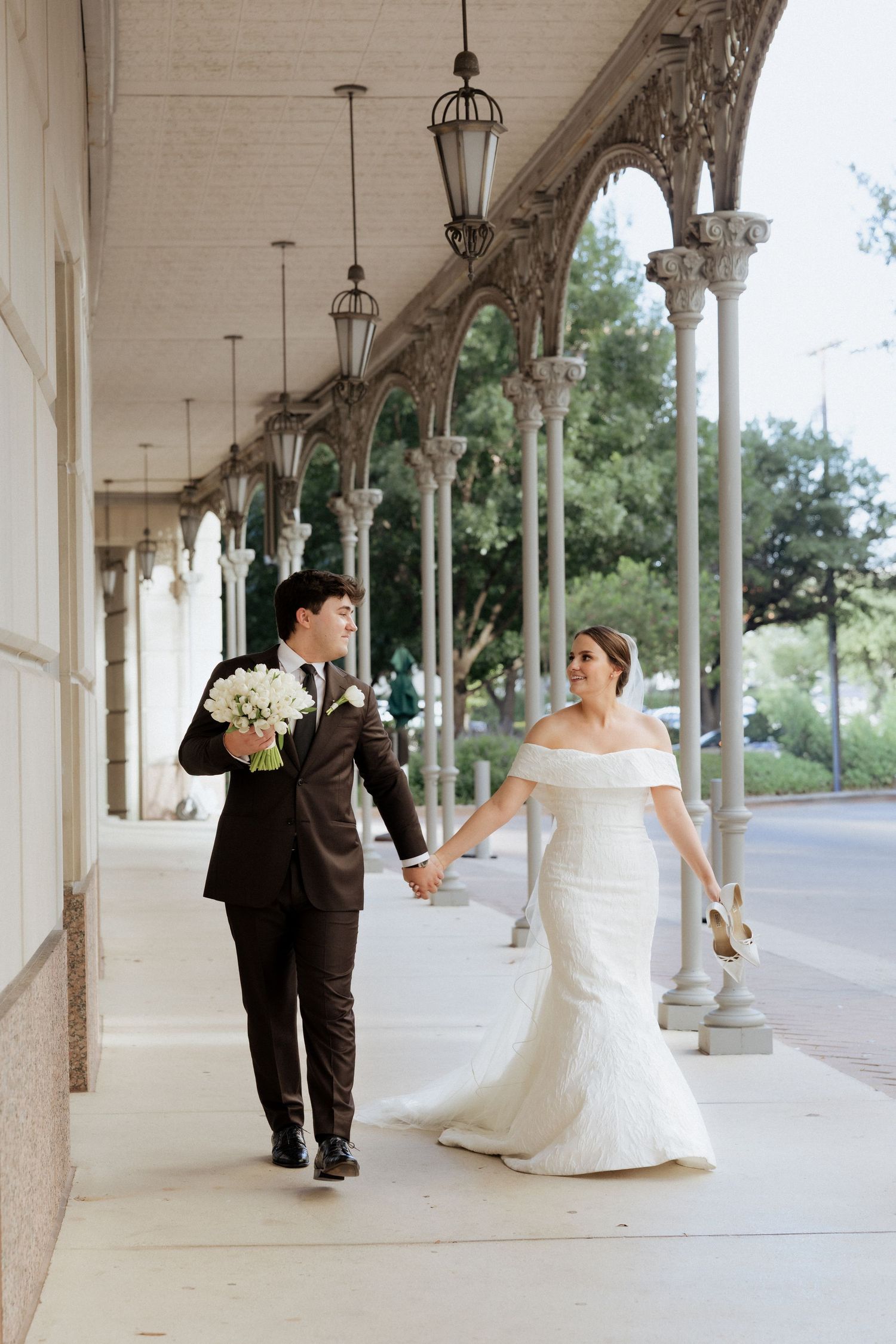 Bride and groom walk hand-in-hand under elegant arched colonnade, she in off-shoulder gown, he in dark suit holding bouquet.
