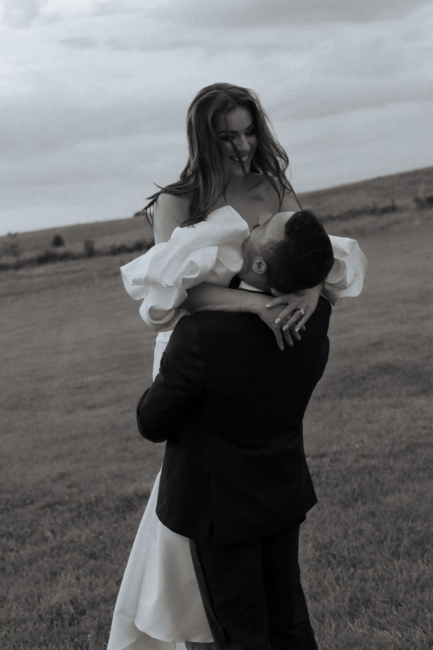 Black and white photo of groom lifting bride in white dress outdoors in a field on their wedding day.