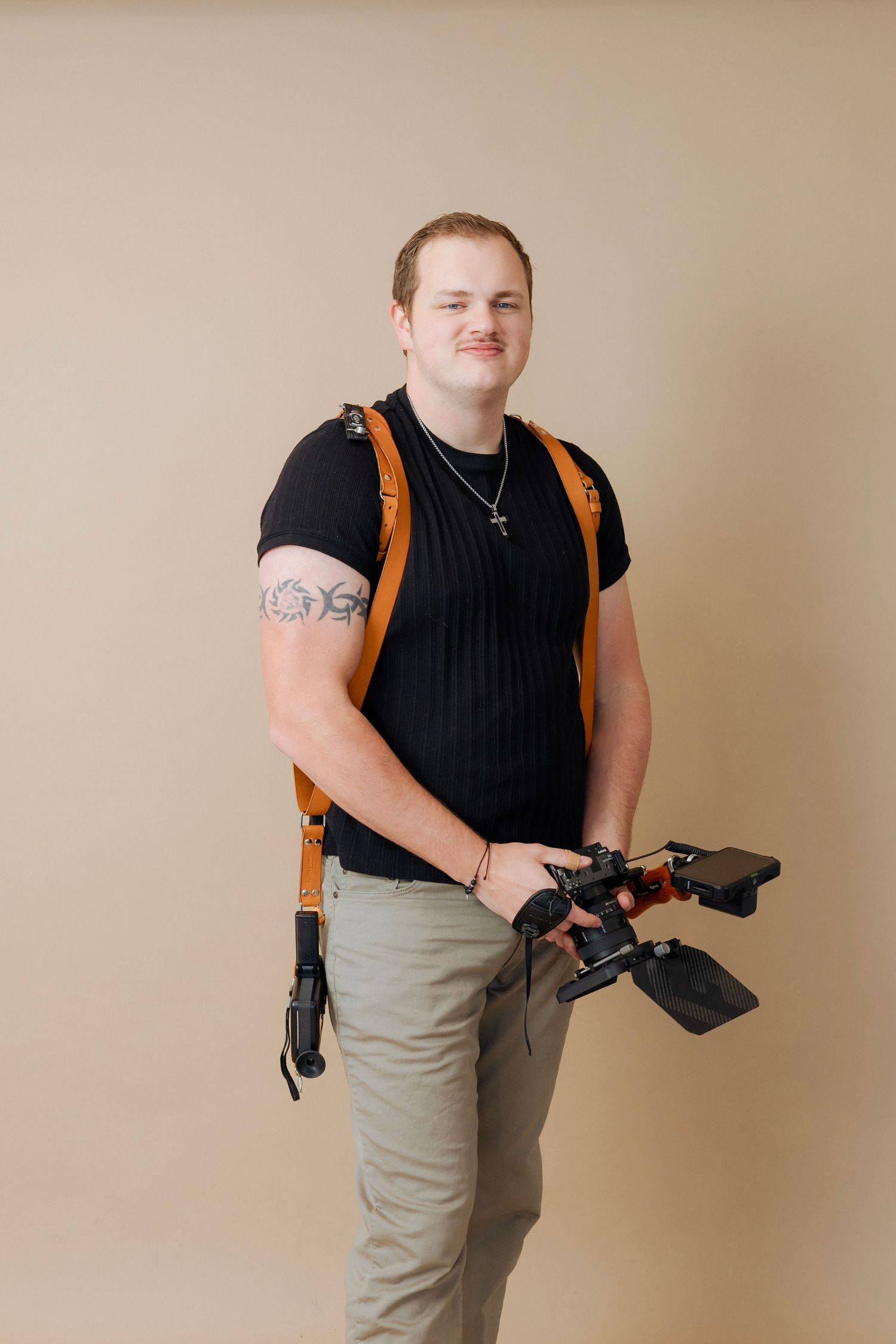 A photographer in a black t-shirt and khaki pants stands holding camera gear with a brown harness against a beige backdrop.