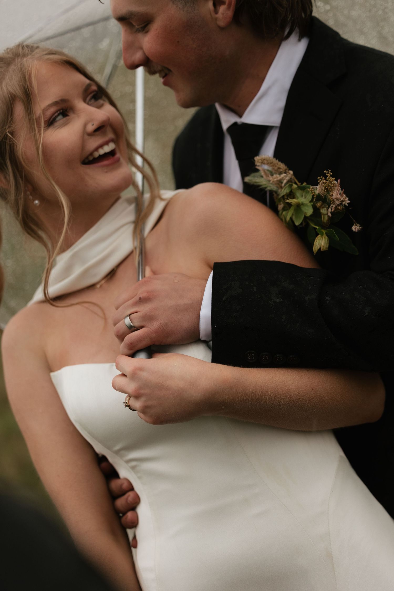 Bride in strapless white gown laughs joyfully as groom in black suit embraces her under a clear umbrella.