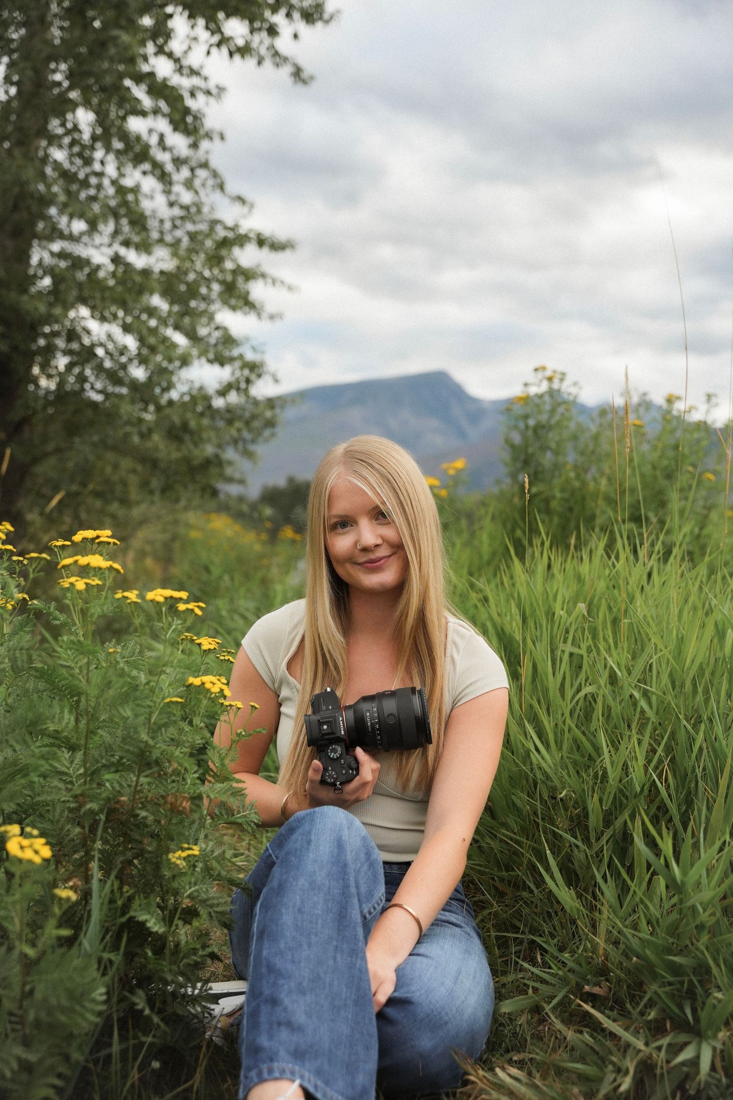 A photographer in denim sits in a meadow of tall grass and yellow wildflowers with mountains in the background.