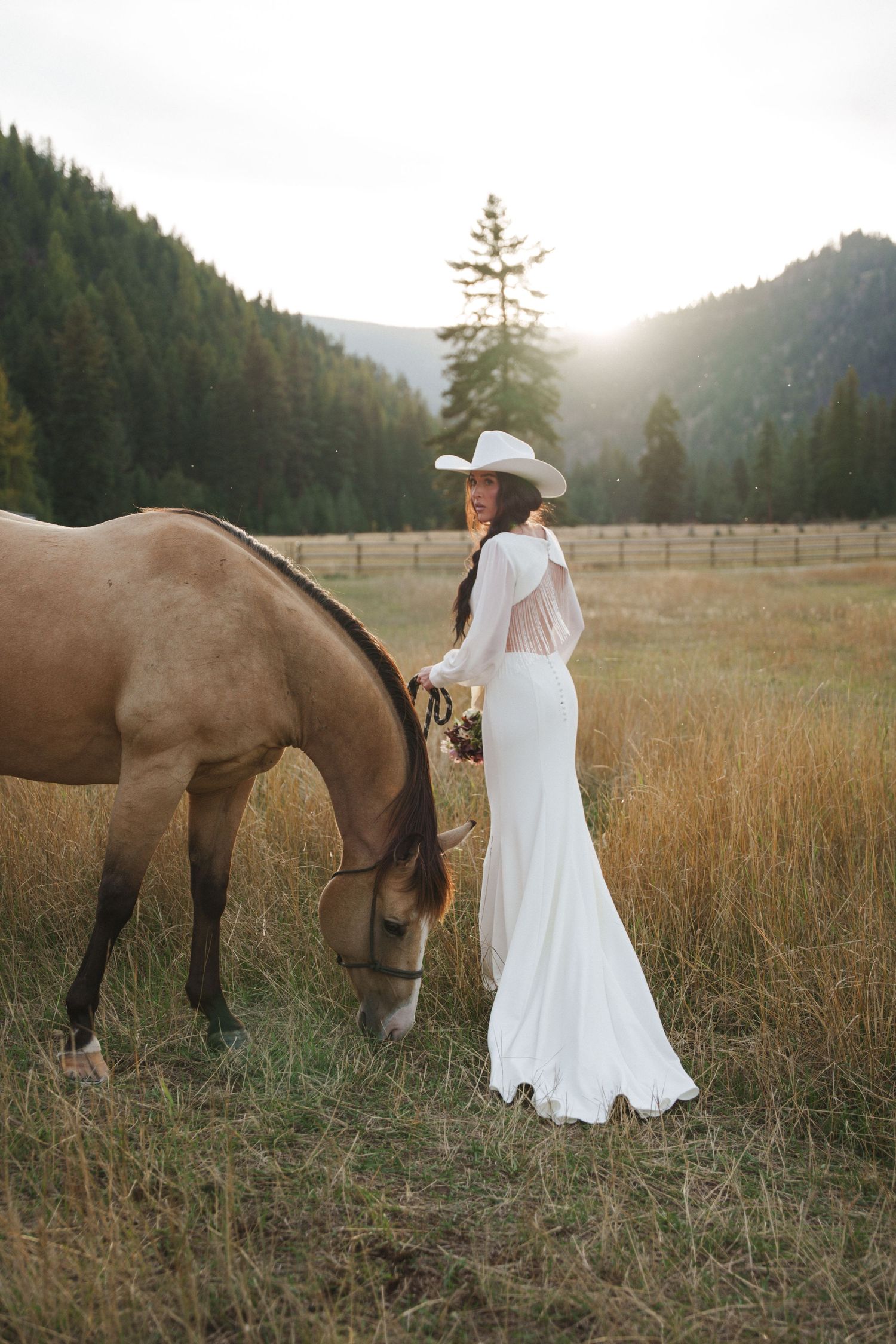 Bride in white gown and cowboy hat stands beside a grazing brown horse in a sunlit mountain meadow.