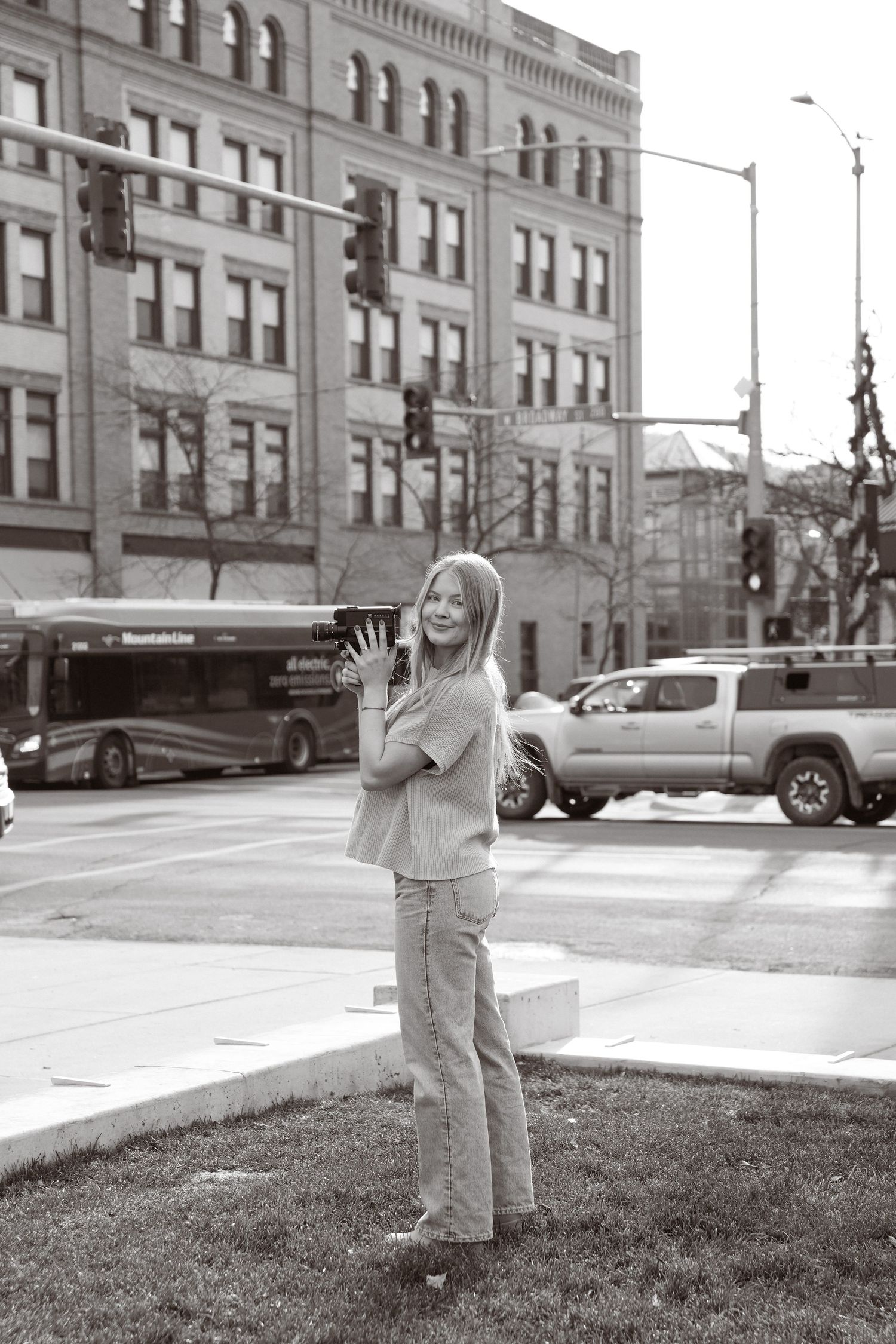 Black and white urban street scene with person standing on grass near city buildings and traffic light.