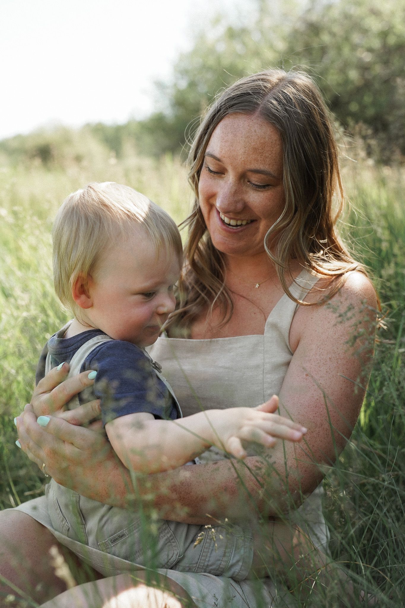 A tender moment between parent and child sitting together in tall grass during a sunny outdoor photo session.