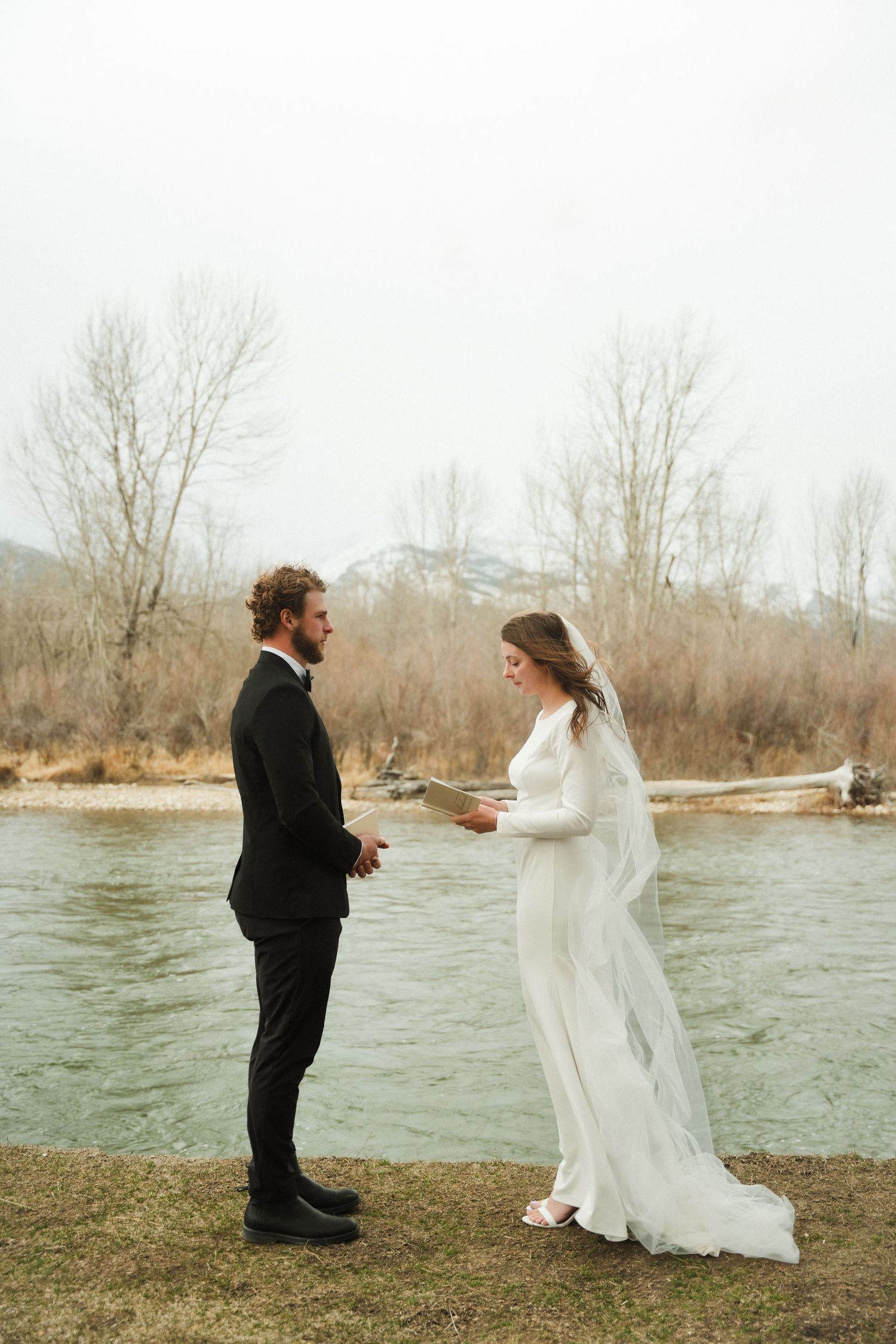 Bride reads vows to groom in white dress and veil by a river, surrounded by bare trees on a cloudy day.