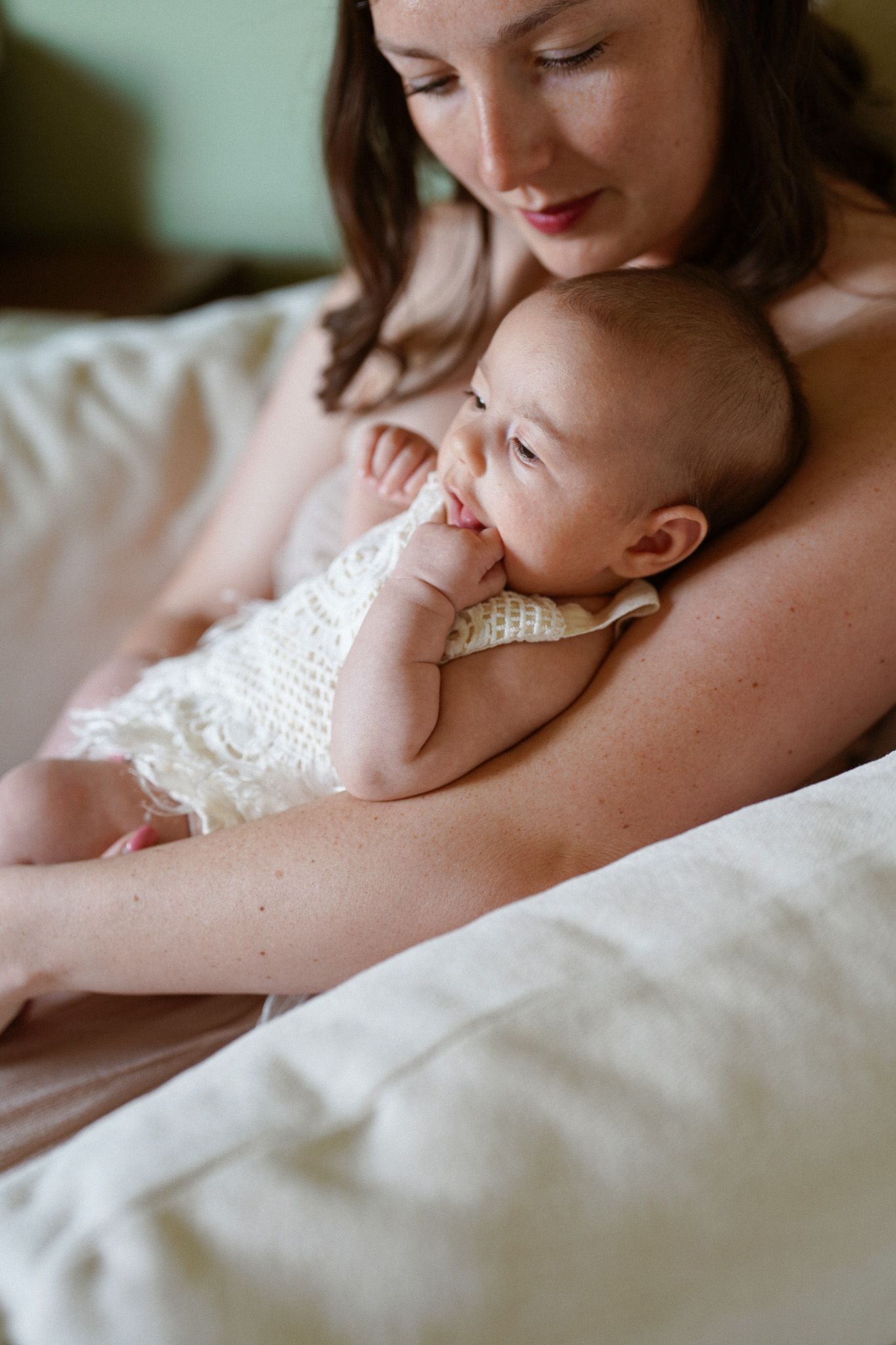 A tender moment of a mother cradling her newborn baby in a white lace outfit on a soft beige bed.