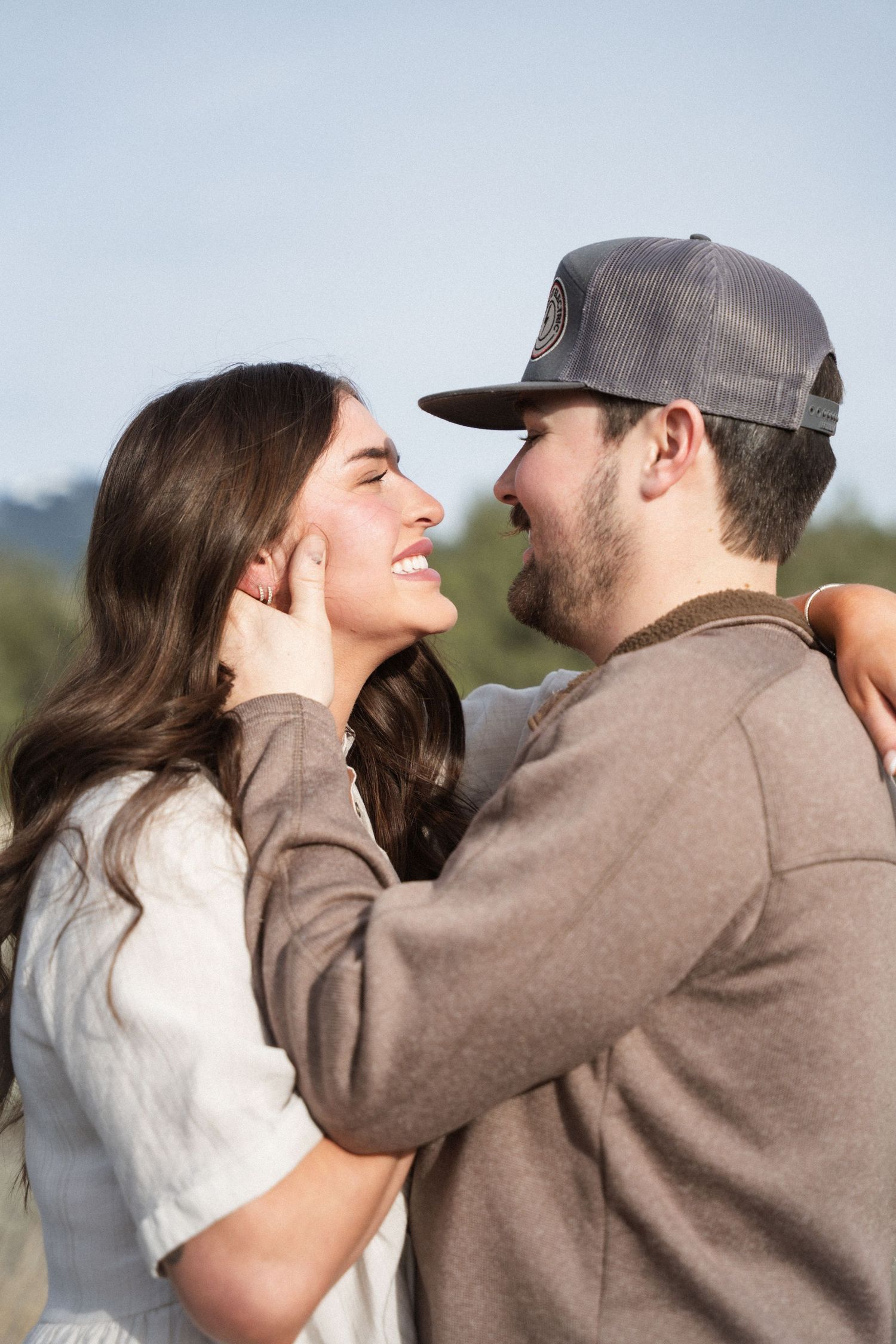 A smiling couple shares a tender moment outdoors, man in gray cap gently holding woman's face.