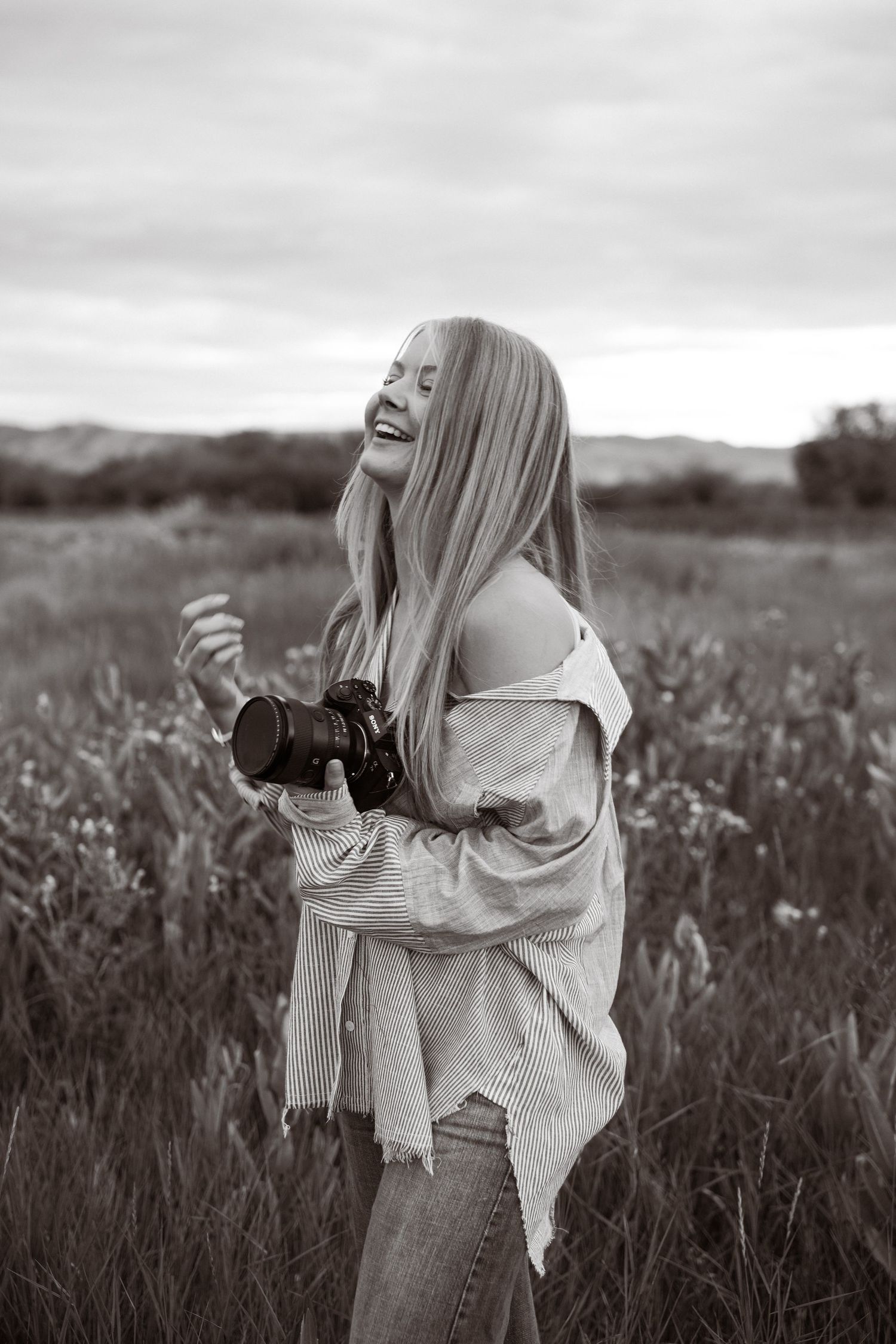 A person in a light sweater holds a cat while standing in a misty field of tall grass at dusk.