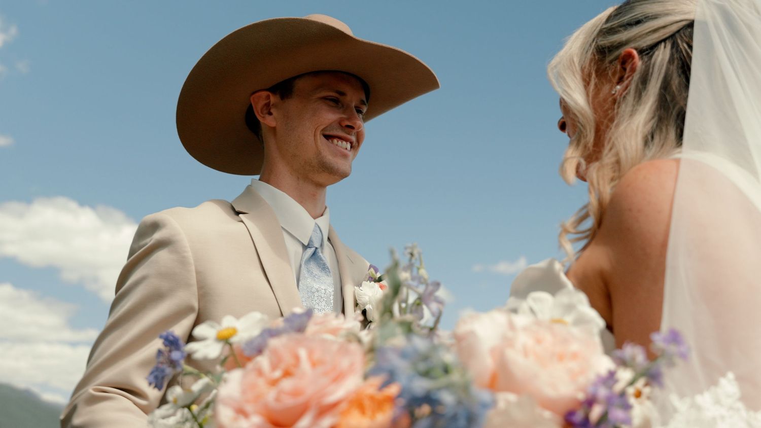 Groom in tan cowboy hat and suit smiles at bride holding colorful bouquet at outdoor wedding.