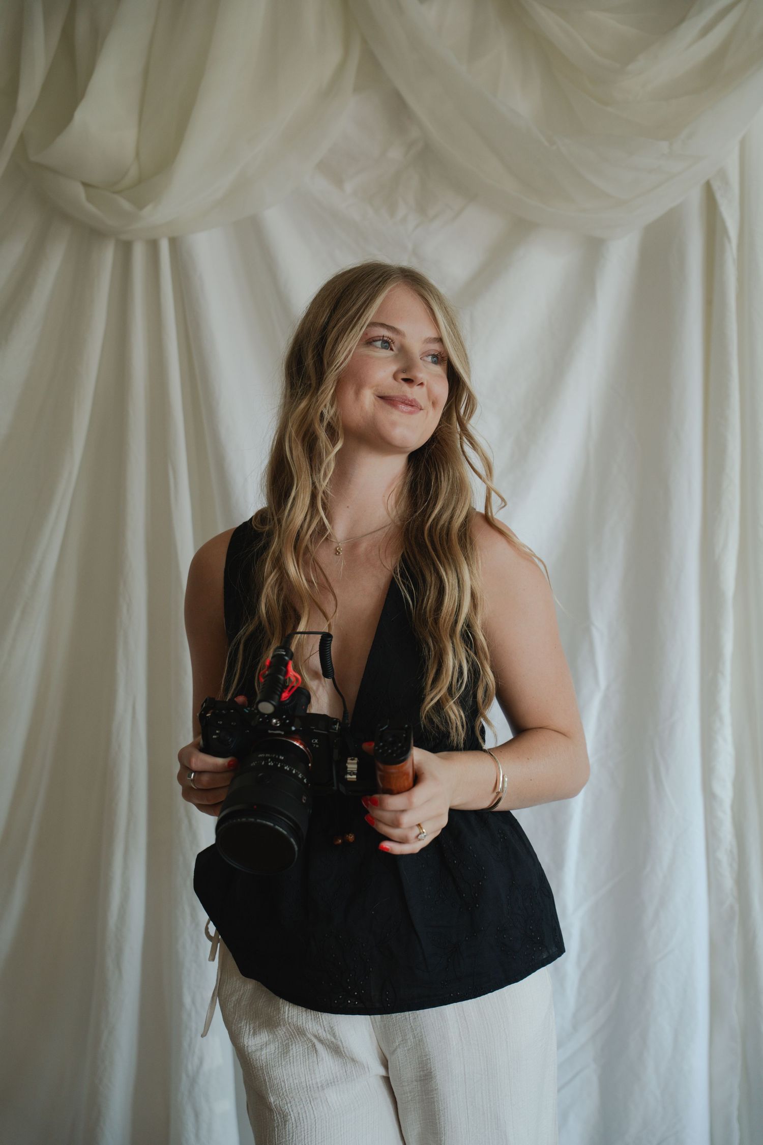 Smiling blonde woman holding a DSLR camera, wearing a black top and white pants against a white draped backdrop.