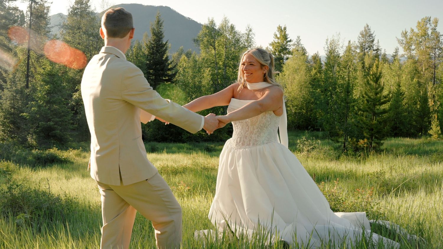 A bride and groom dance together in a sunlit mountain meadow surrounded by evergreen trees on their wedding day.