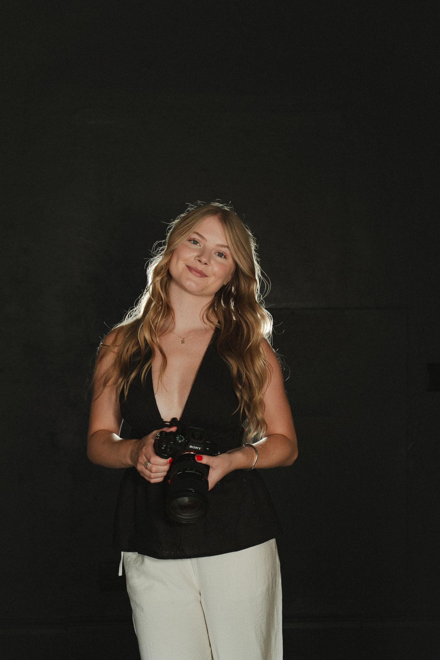 Smiling woman with long wavy hair holding a camera, wearing a black top and white pants against a dark background.