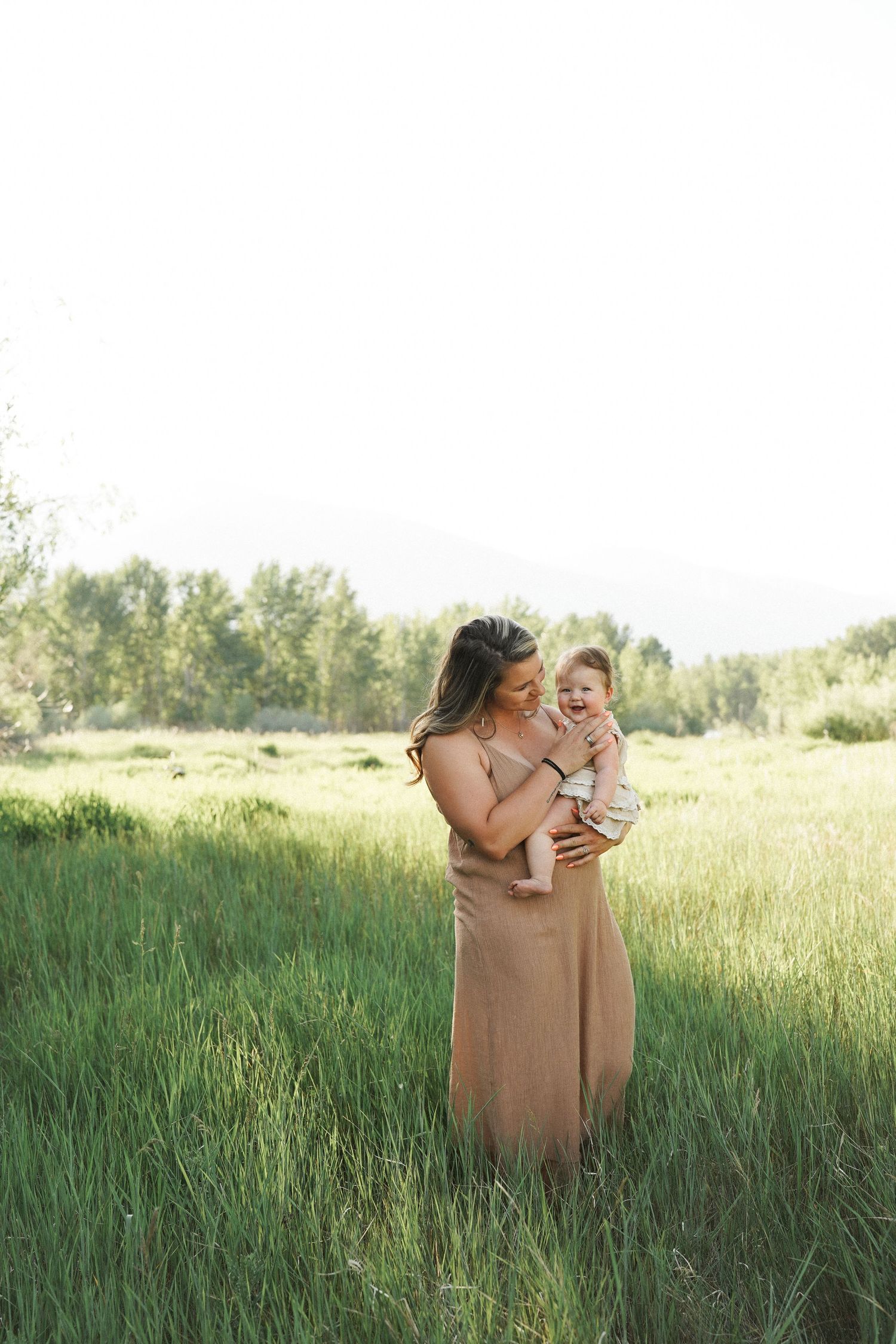 A mother in a tan dress holds her baby in a lush green field, sharing a tender moment outdoors.