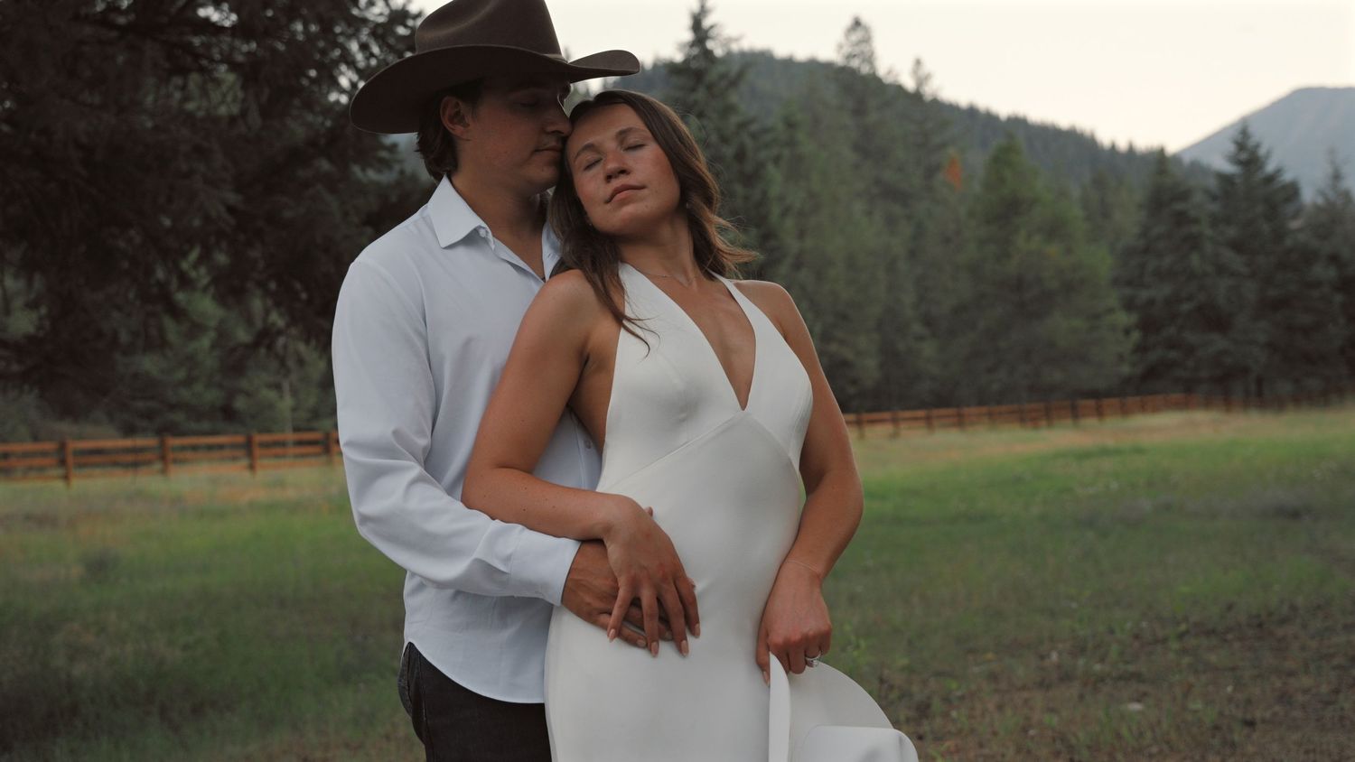 Romantic couple embraces in a scenic mountain meadow at sunset with rustic wooden fence in background.