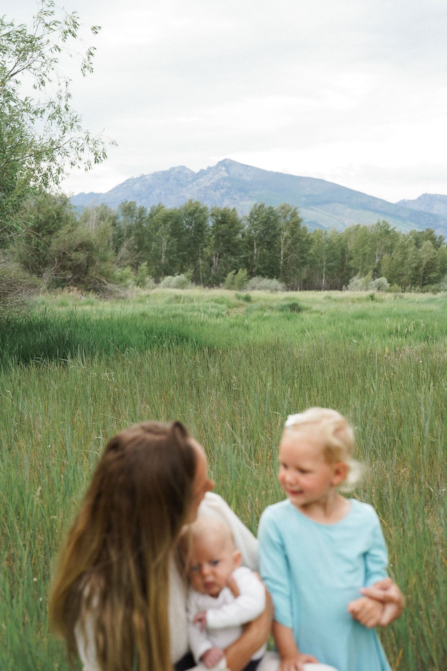 A mother holds her baby while her young daughter in a teal dress plays in a lush green meadow with mountains behind.