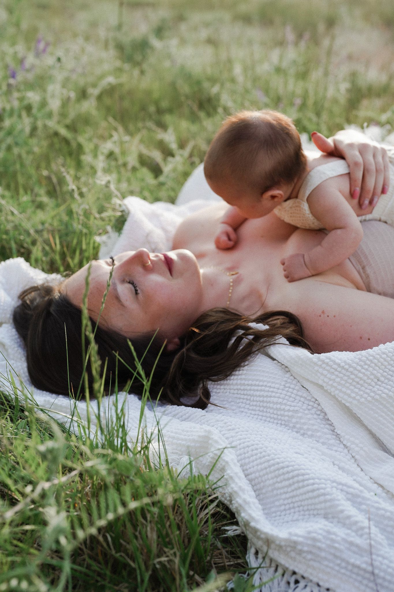 A mother lies on a blanket in a green meadow, cradling her newborn baby on her chest in a tender moment.