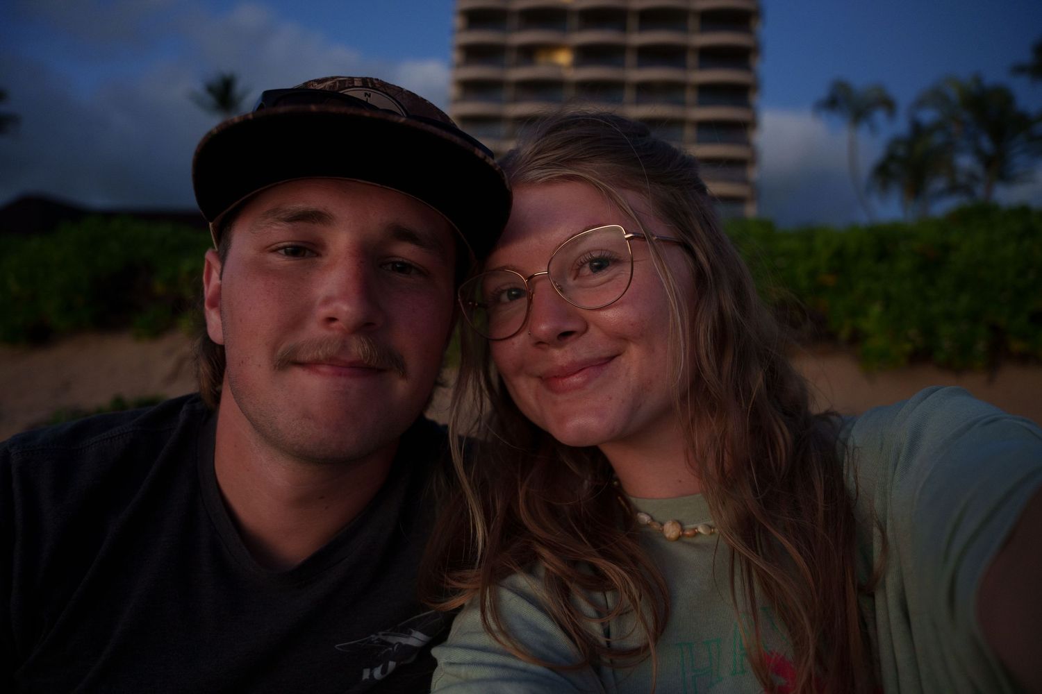 A young couple smiles together for a selfie on a beach at dusk with a tall building and trees in the background.