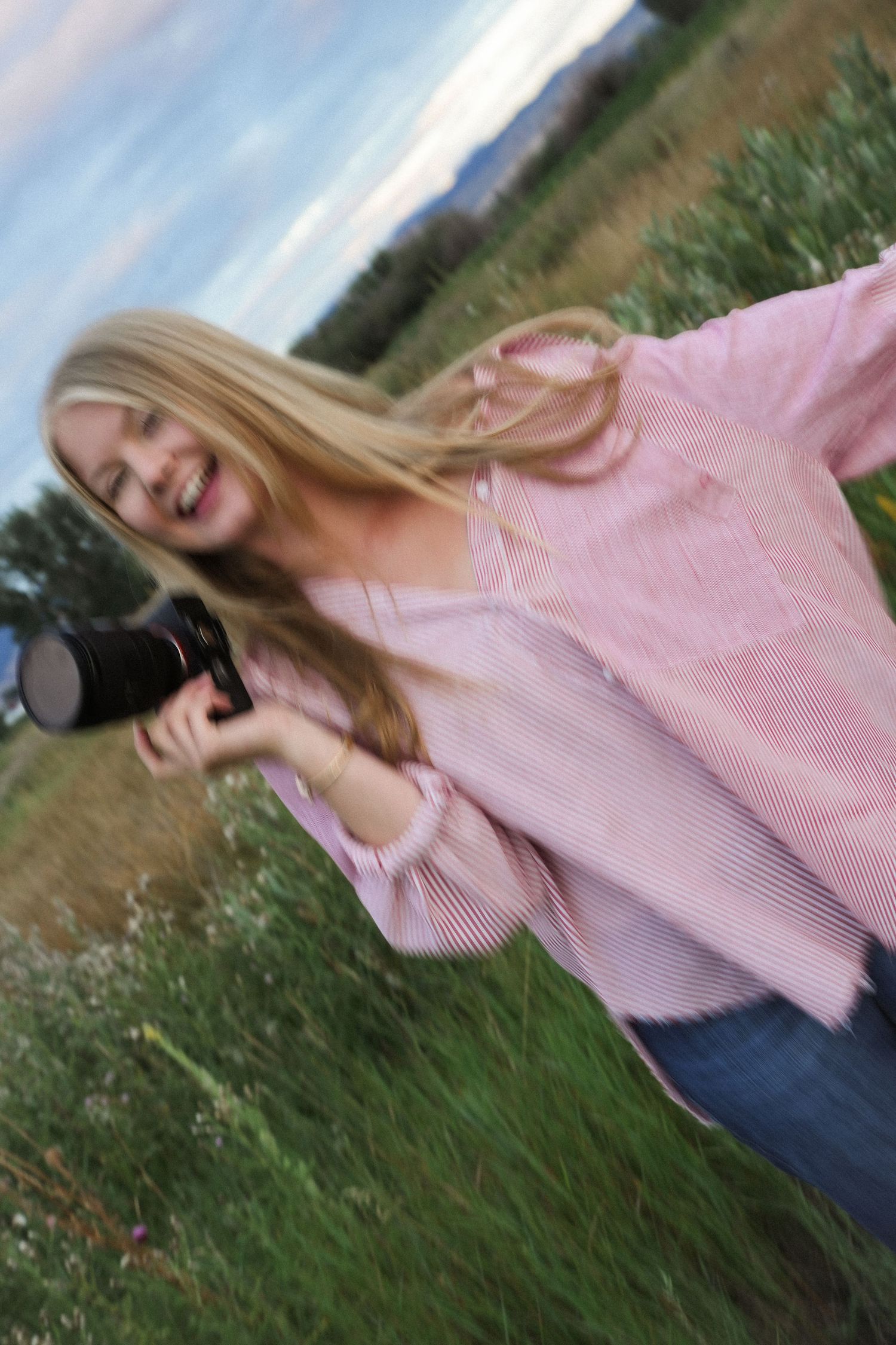 A person in a pink striped sweater and jeans poses outdoors against a natural landscape background.
