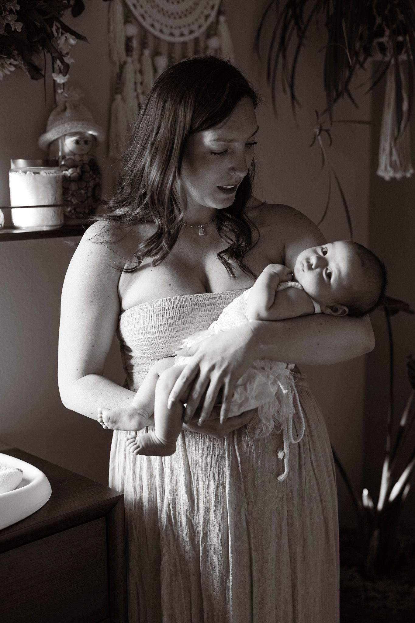 Black and white photo of a young mother in a white dress tenderly holding her newborn baby in a nursery room.