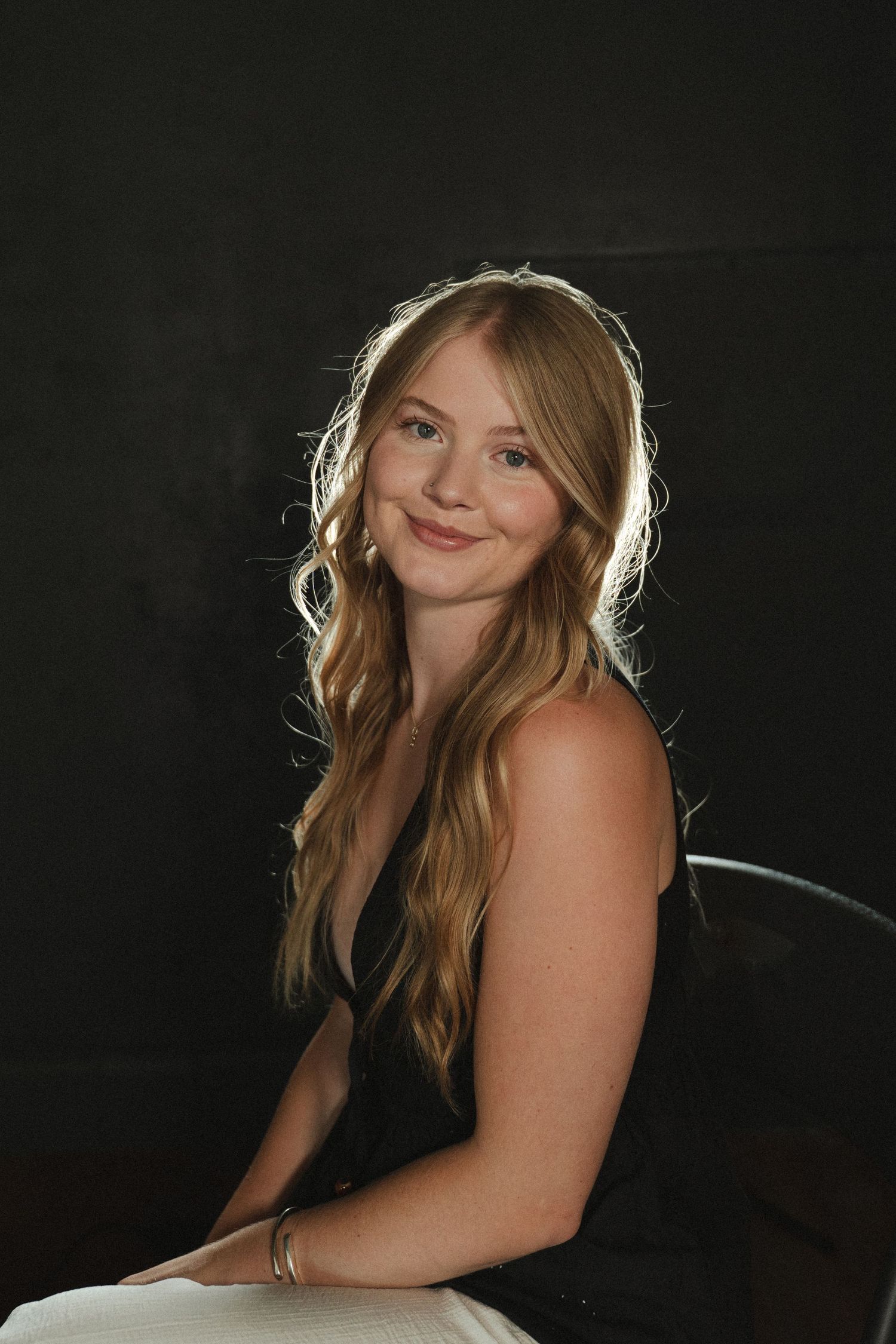 Smiling young woman with long wavy blonde hair in a black sleeveless top sits against a dark background in a softly lit portrait.