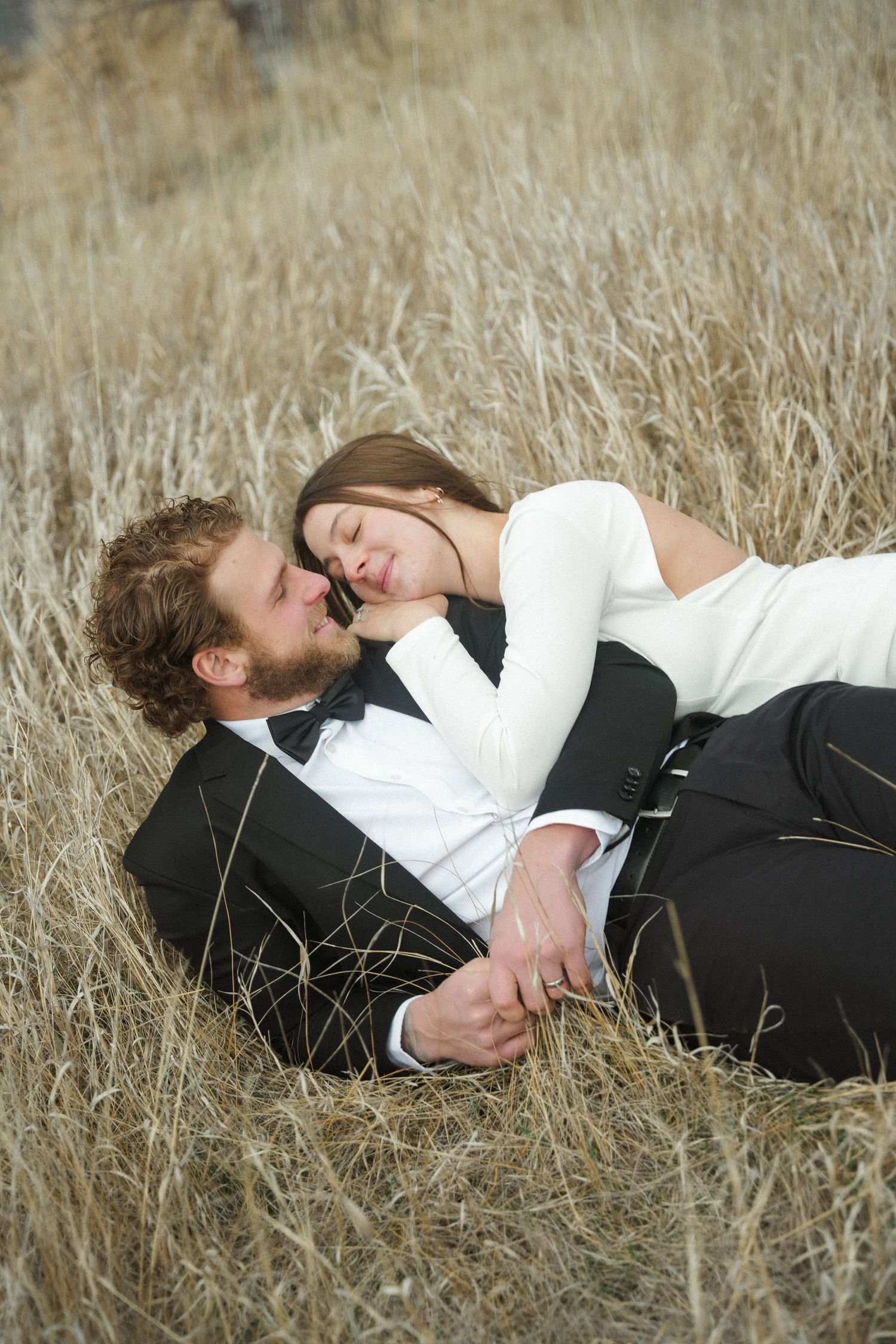 A romantic couple in formal attire lying together in a golden wheat field, sharing an intimate moment.