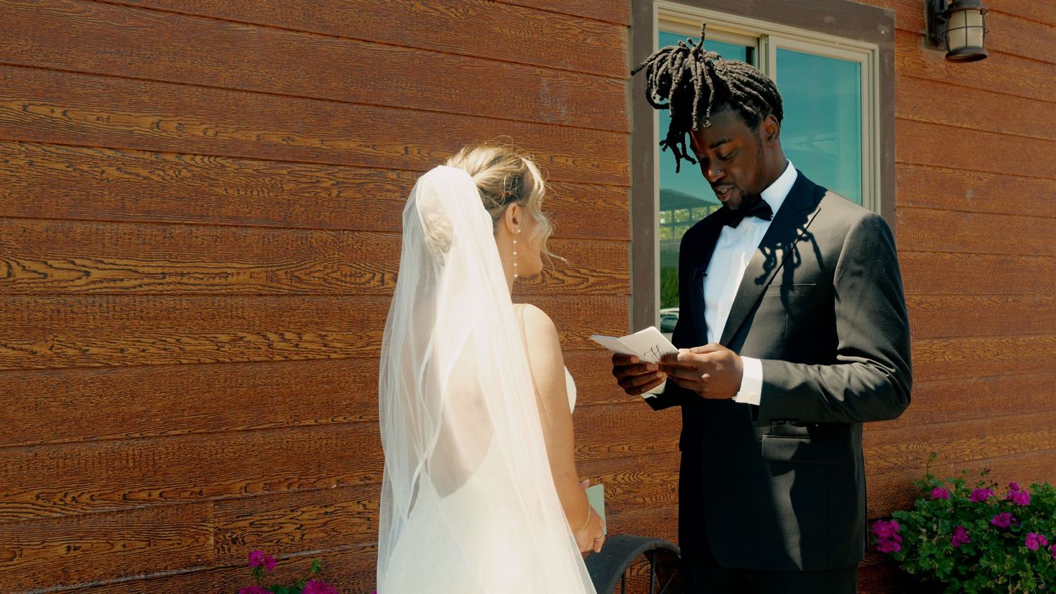 A couple exchanges vows during an outdoor wedding ceremony against a wooden wall with a turquoise door.