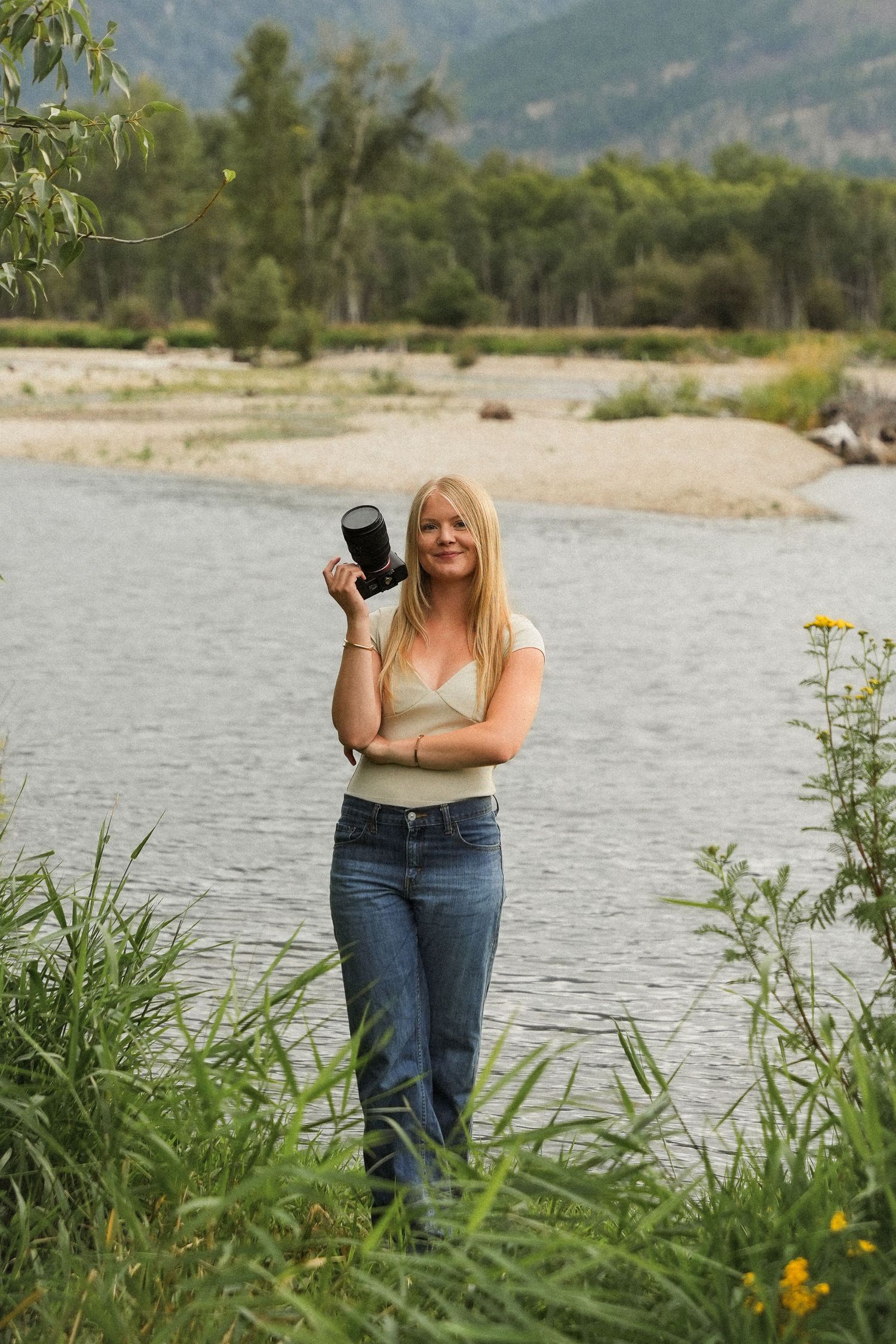 Person standing by a lake with mountains and trees in the background, holding a camera and taking photos.