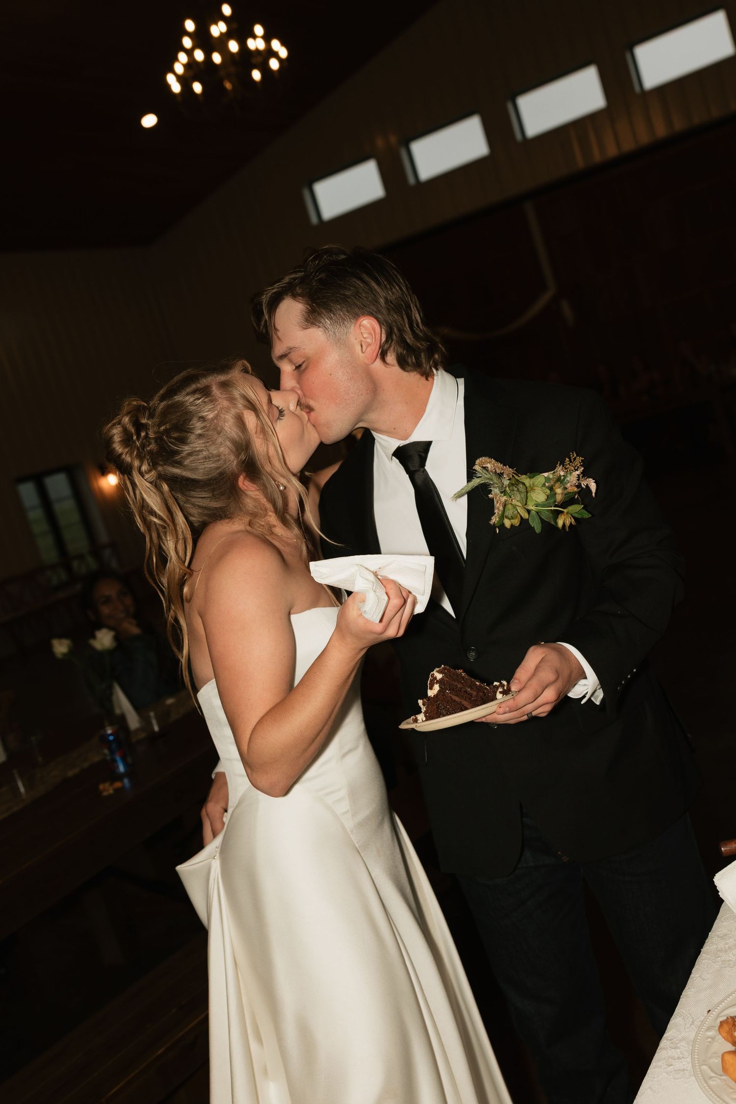 Bride and groom share a kiss while holding wedding cake slices at their indoor reception venue.