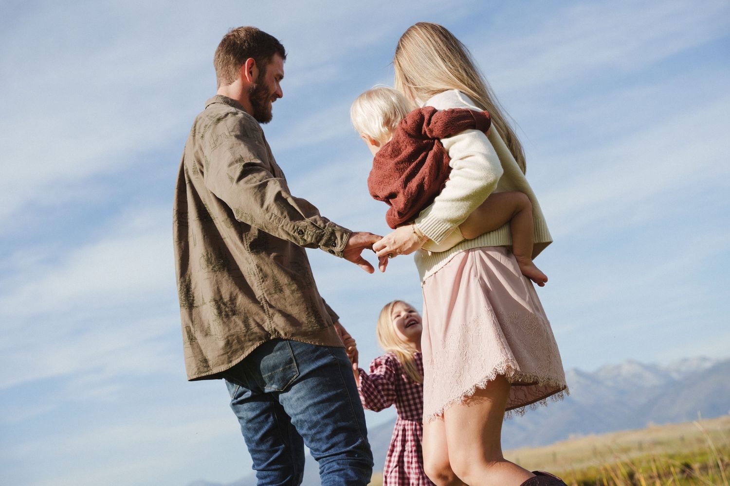 Family of four enjoying outdoor time together, with parents holding hands with young children in a scenic mountain field.