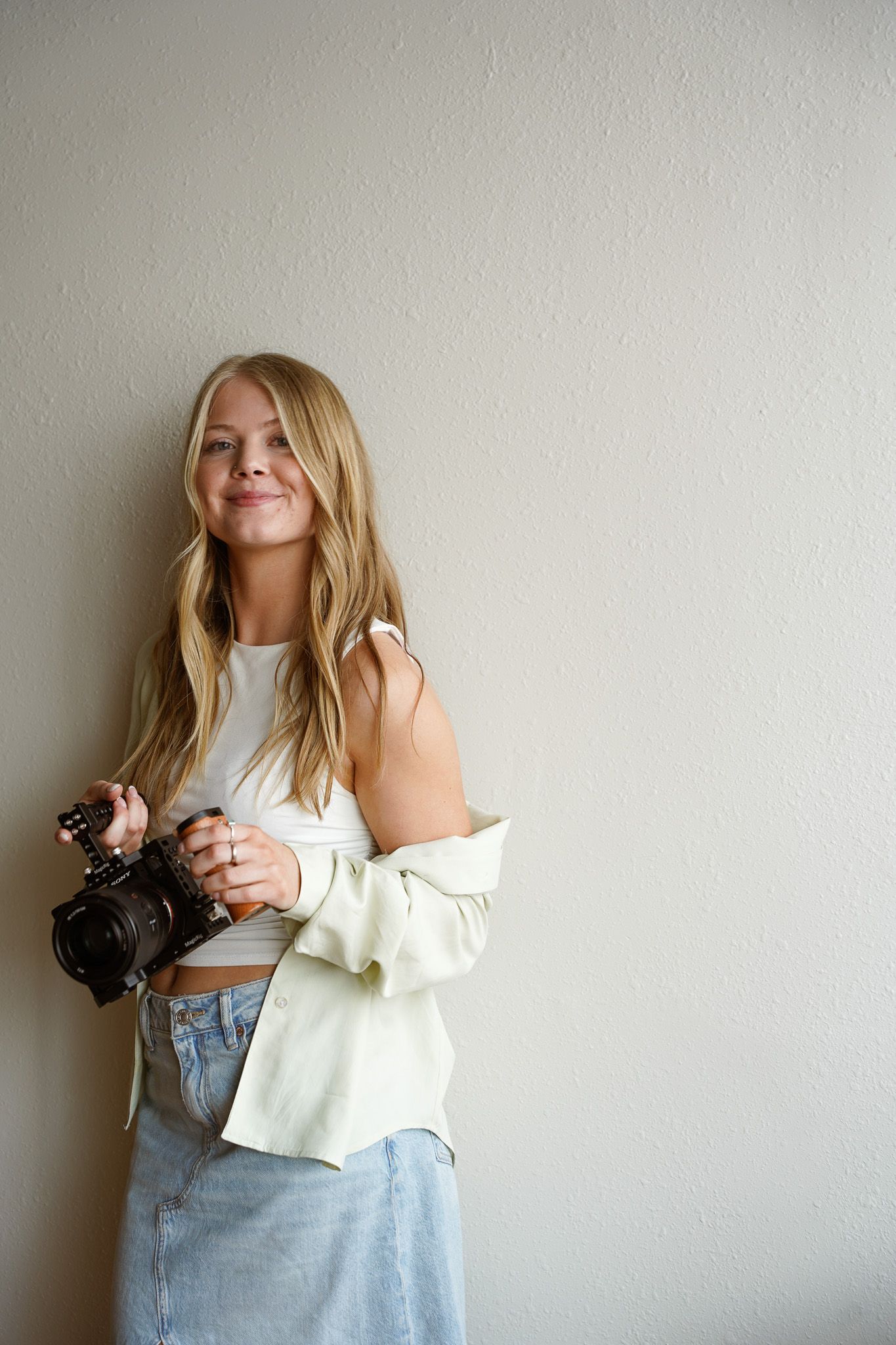 Young blonde woman smiling and holding a camera, wearing a white crop top and light blue jeans against a neutral wall.