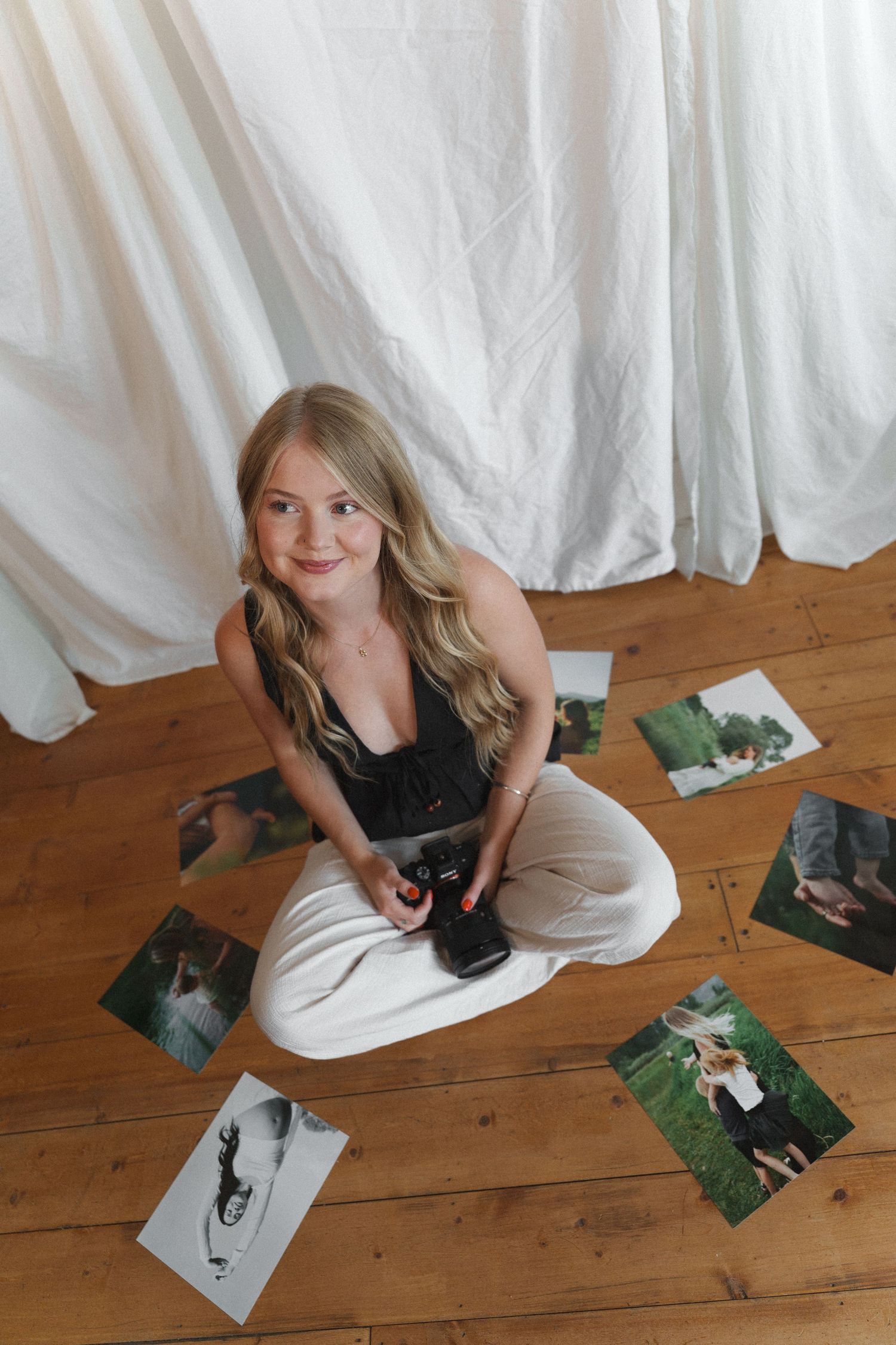 Young blonde woman sitting cross-legged on wooden floor holding camera, surrounded by printed photos, against white fabric backdrop.