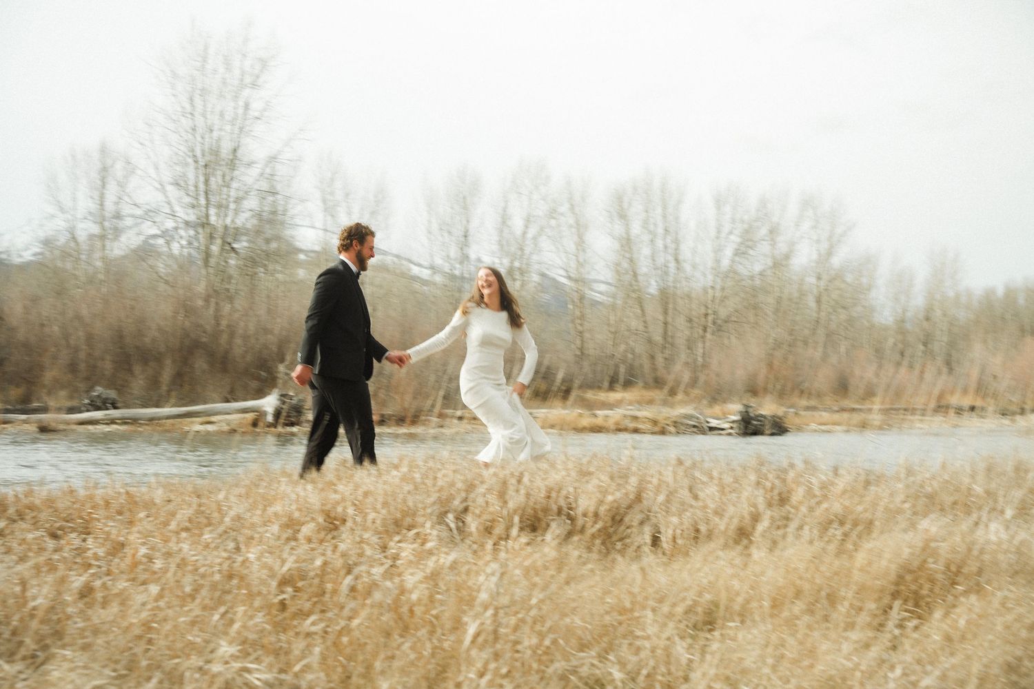Bride and groom holding hands walking through golden fields near a river on their wedding day.