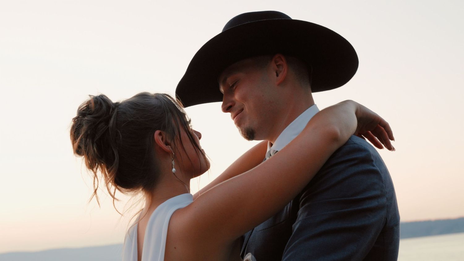 A couple in formal attire embrace during sunset by the water, with the person wearing a black hat and suit.