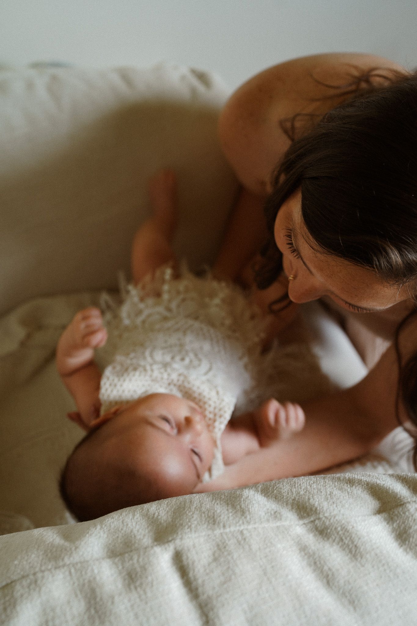 A mother tenderly gazes at her sleeping newborn baby wearing a white knit bonnet on a soft blanket.