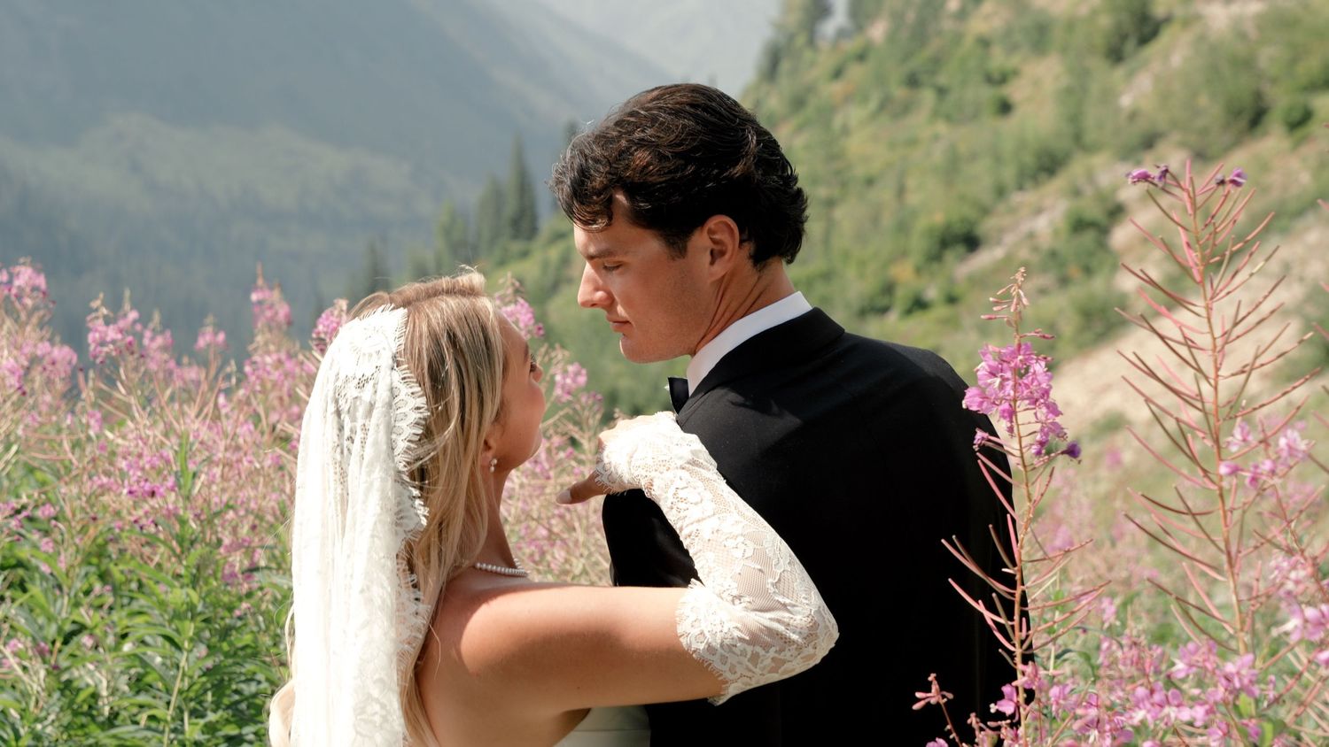 Bride and groom share romantic moment outdoors surrounded by pink wildflowers with mountain scenery backdrop.