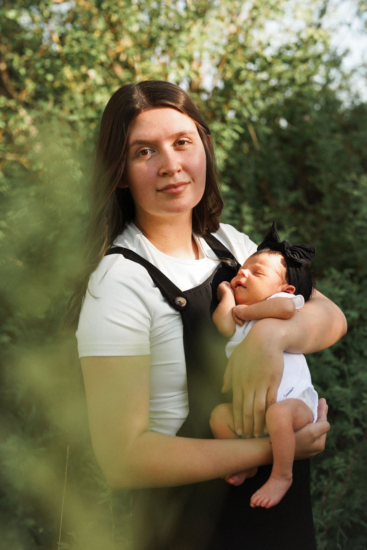 A young mother tenderly holds her sleeping newborn baby outdoors, surrounded by lush green foliage.