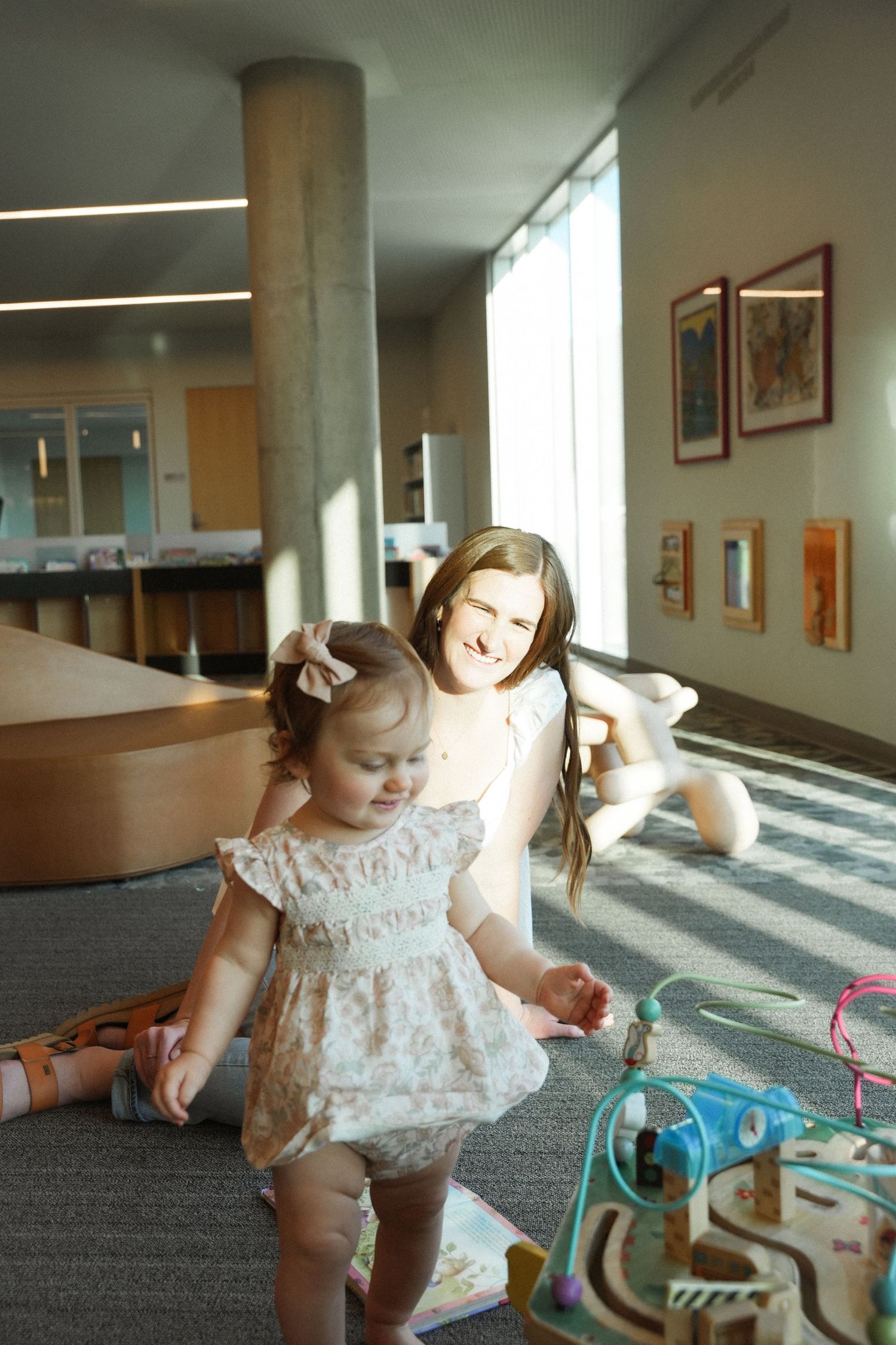 Two young girls playing with toys on carpet in a bright, modern building interior with large windows.