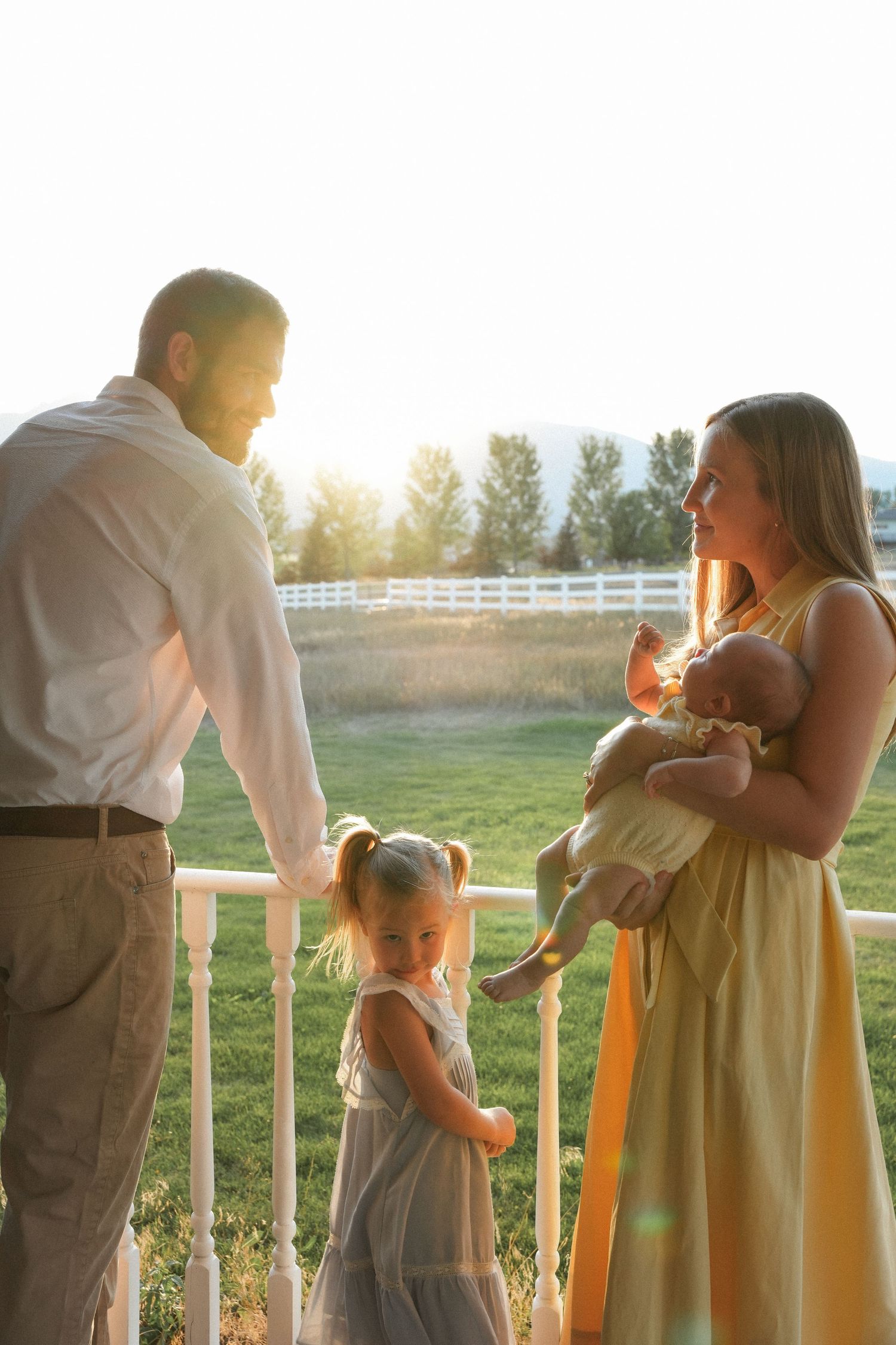 Family stands on porch at sunset with white fence and trees in background during golden hour.